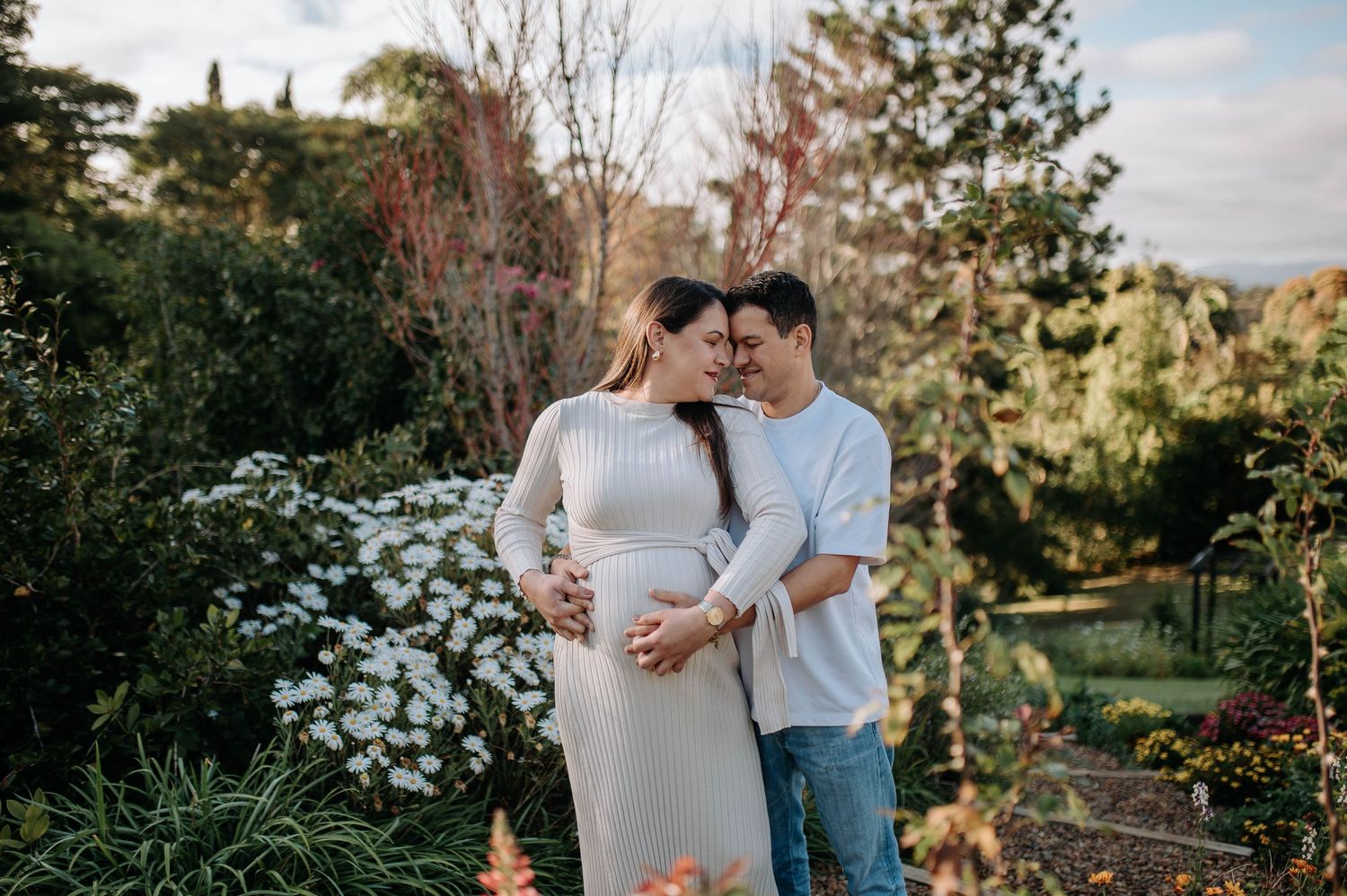 Pregnant couple embracing tenderly in a lush garden, woman in grey dress cradling baby bump, surrounded by white flowers.