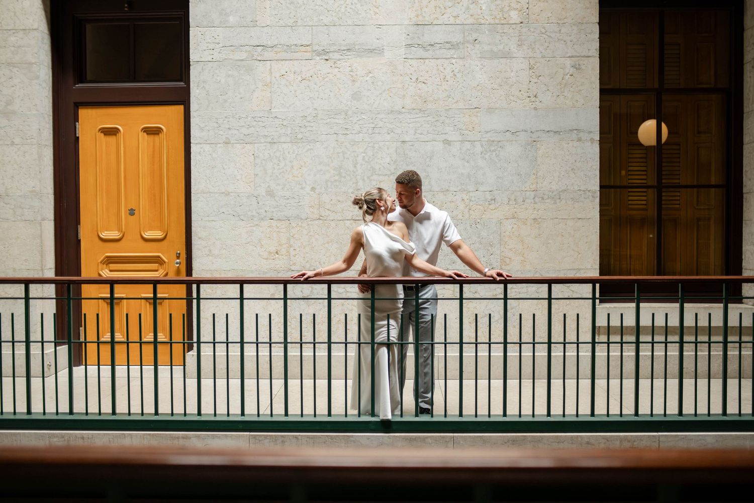 Couple sharing a romantic moment leaning on railing of indoor balcony near yellow door.