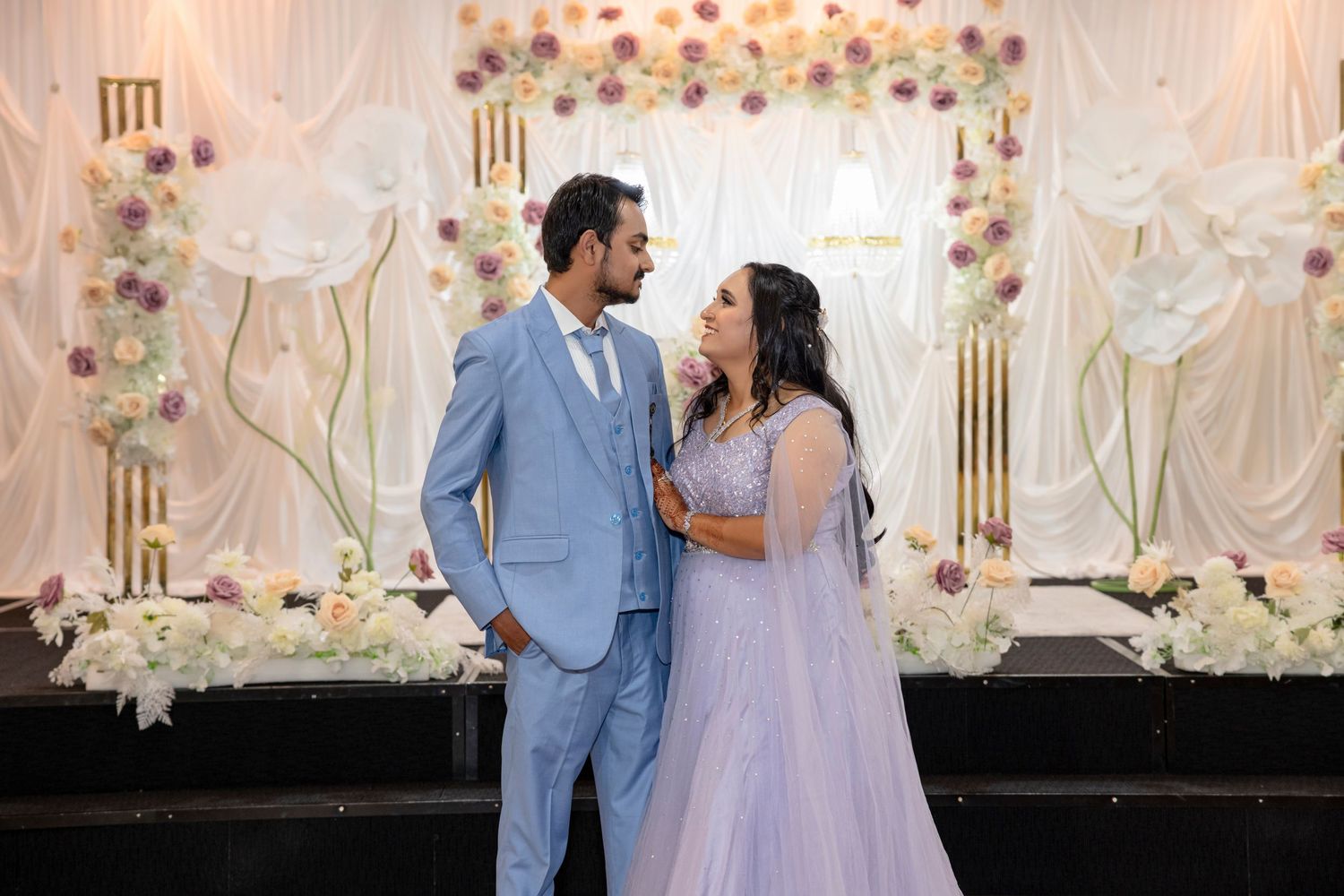 Couple in matching blue and lavender outfits posing in front of a floral backdrop at a wedding celebration.