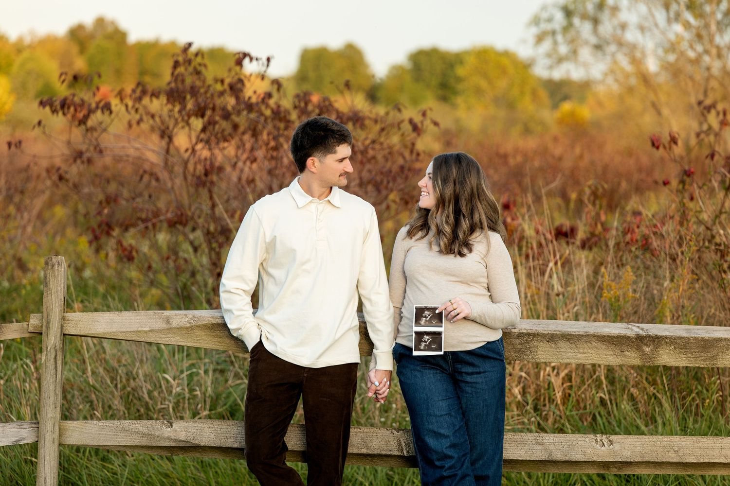 Couple holding hands by a wooden fence, sharing a smile, holding an ultrasound photo outdoors in autumn.