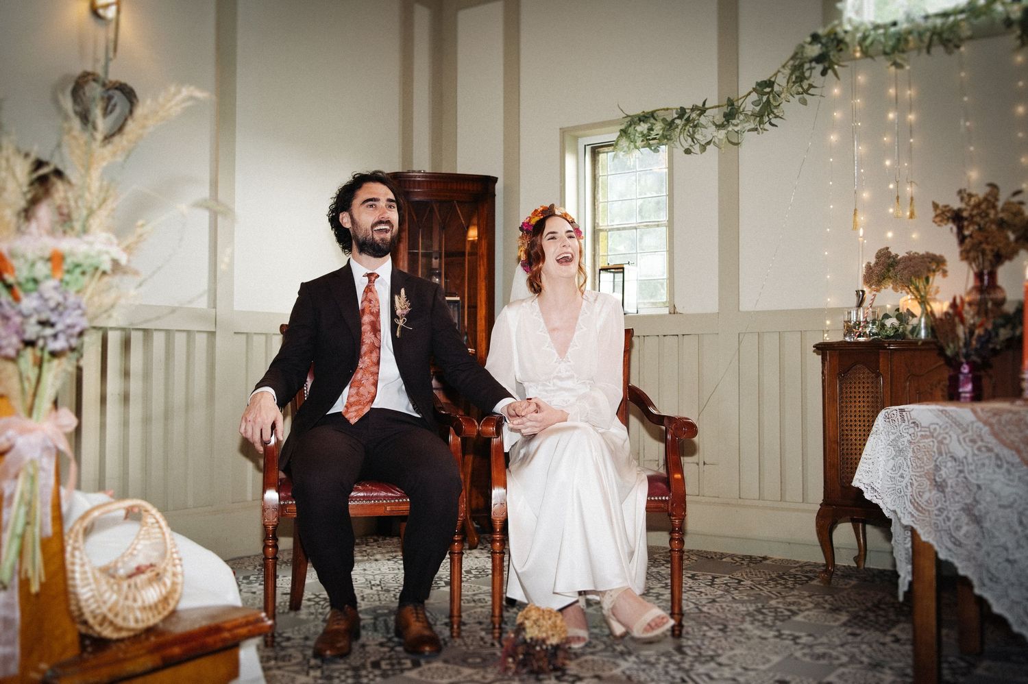 Bride and groom sitting in vintage chairs laughing joyfully during an elegant bohemian indoor wedding ceremony.