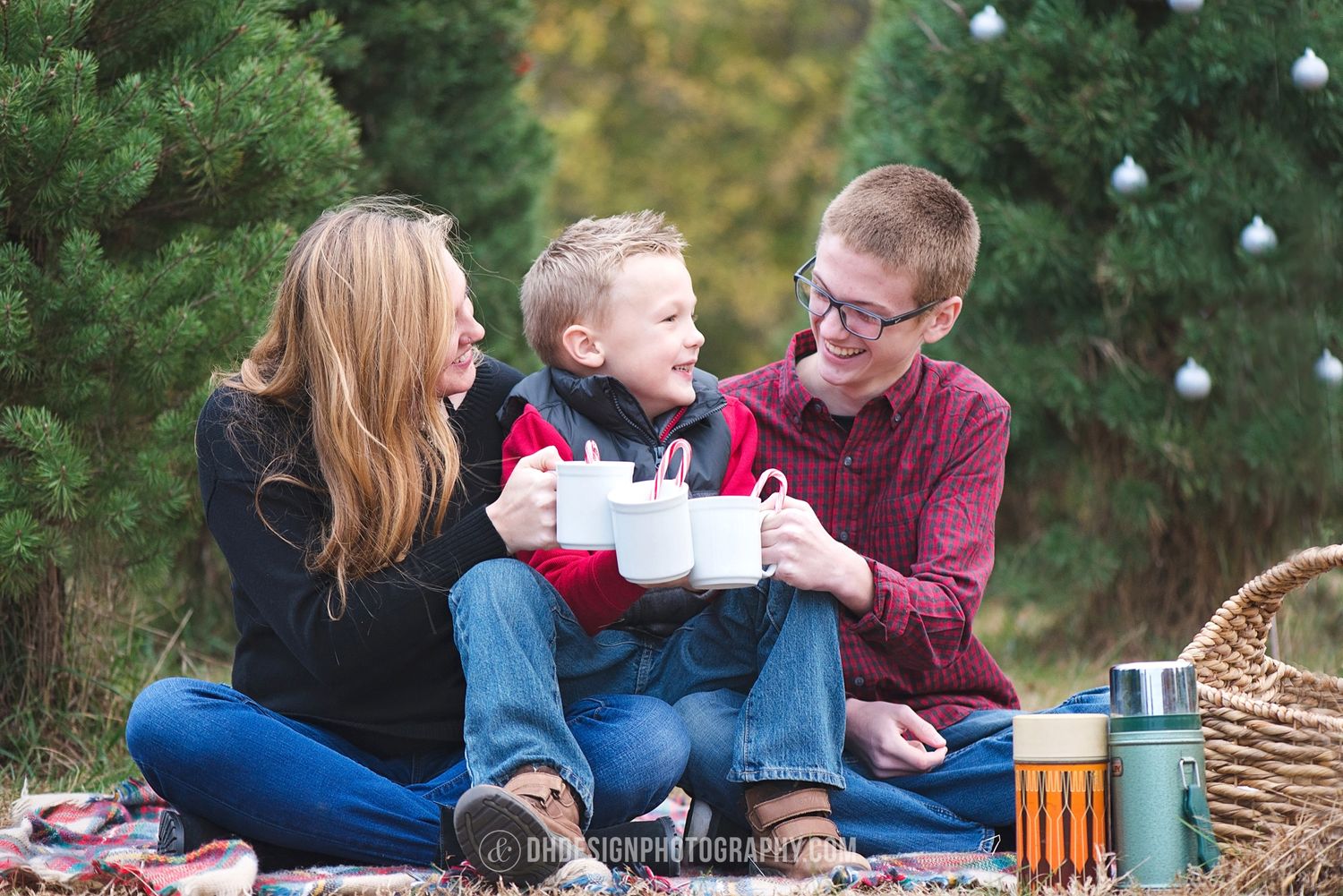 Family of three sitting outdoors on a blanket, smiling and toasting with mugs near Christmas trees.