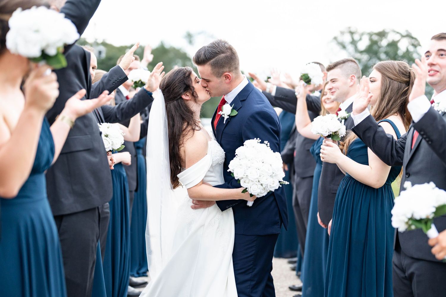 Bride and groom share a kiss surrounded by wedding party in navy dresses and dark suits holding white flowers.