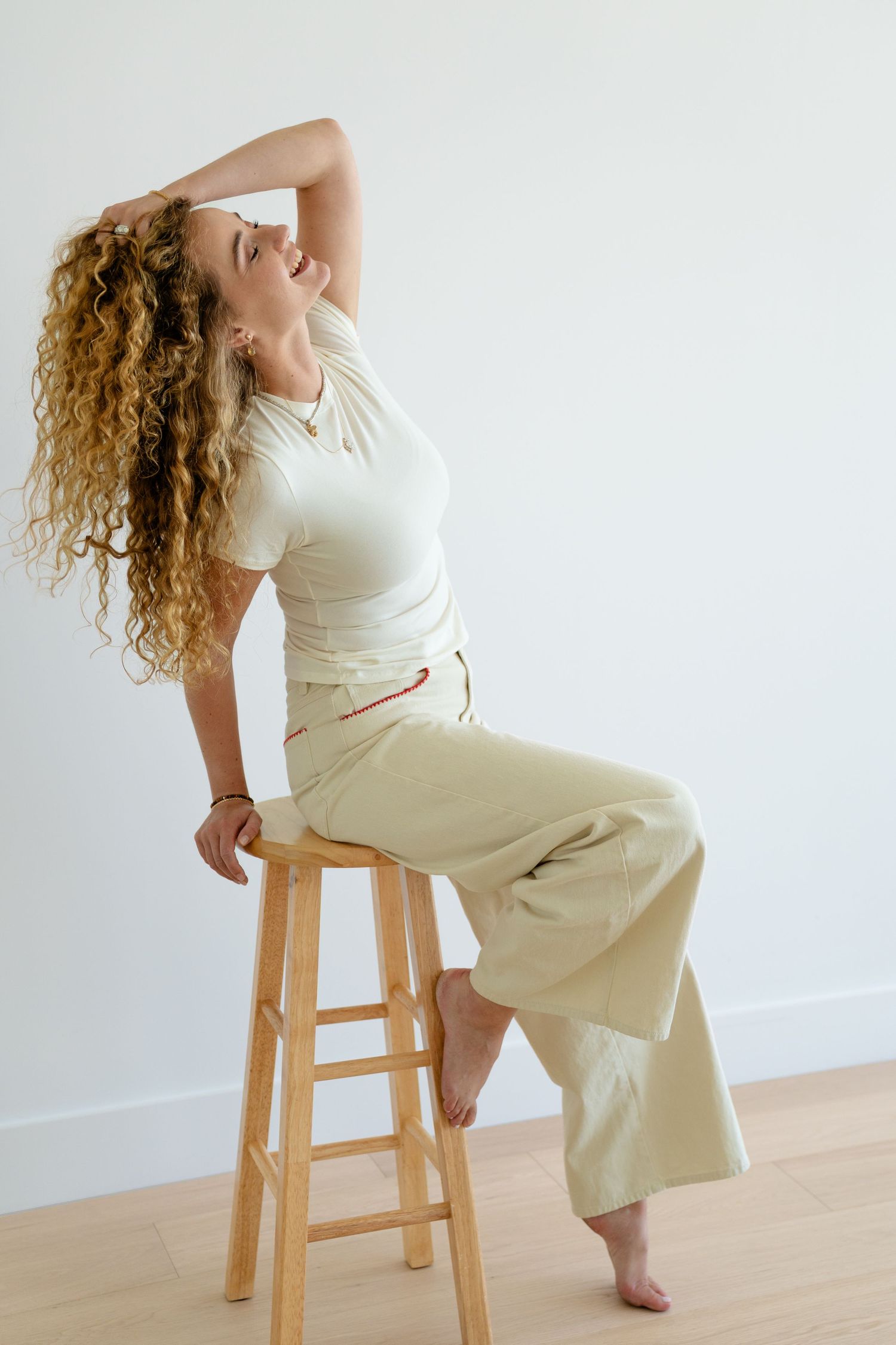 Woman with curly hair sits on wooden stool wearing white tee and wide-leg khaki pants, posing gracefully.