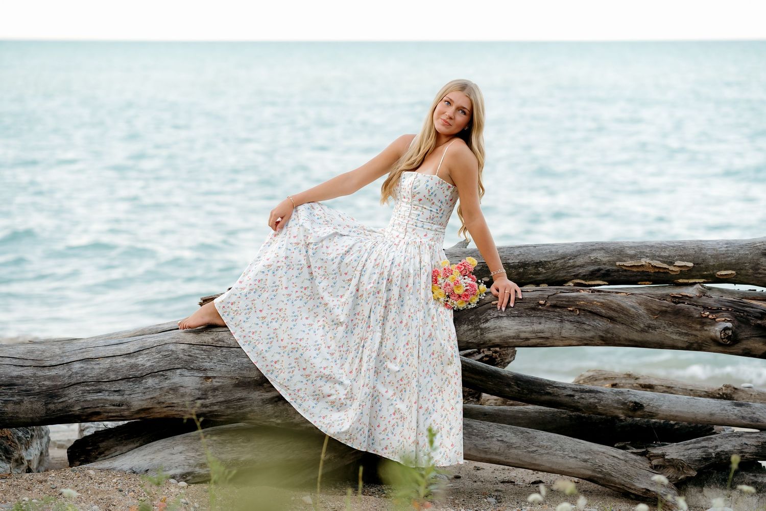 Blonde woman in floral white midi dress posing on rocky beach shoreline with lush green trees in background.