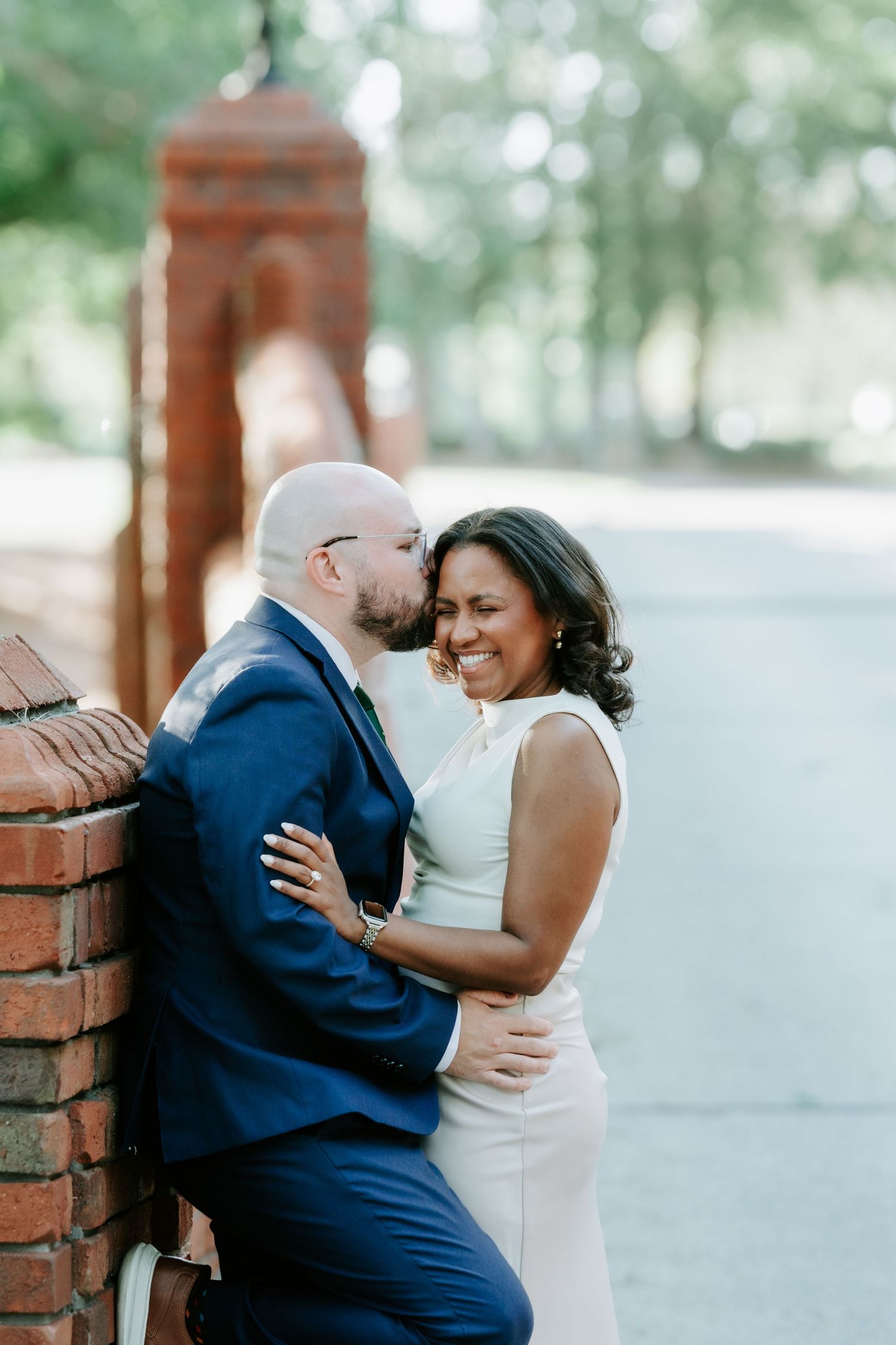 A couple shares a romantic moment by a brick wall, the man kissing her cheek as she smiles joyfully in wedding attire.