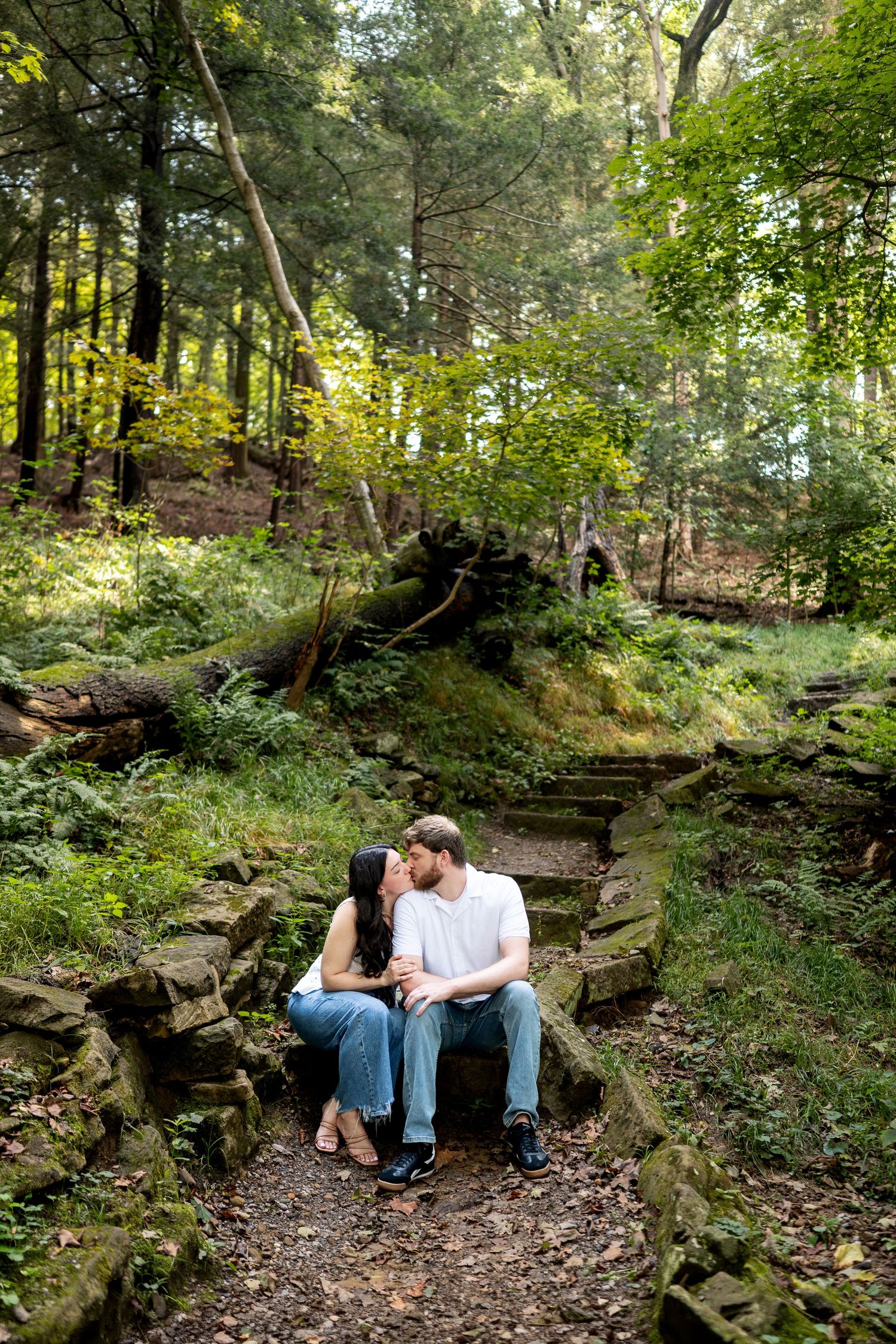A couple shares a kiss while sitting on stone steps surrounded by lush green forest foliage.