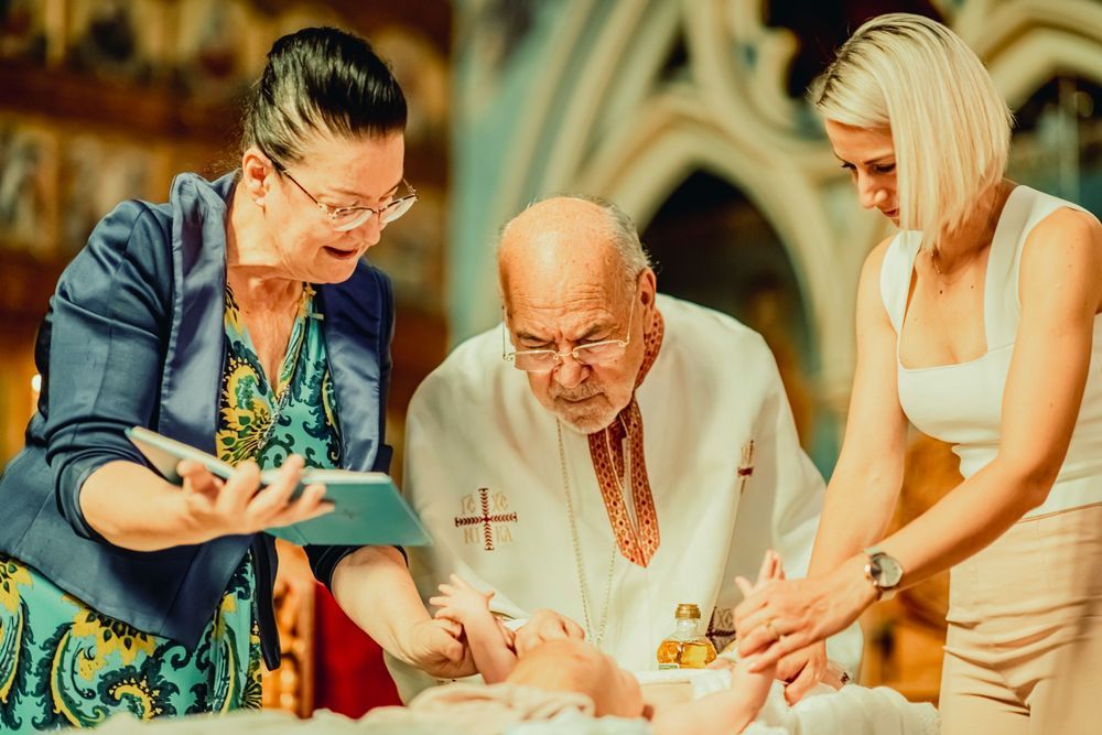 Priest performing a baby baptism ceremony with two women assisting in a decorated church.