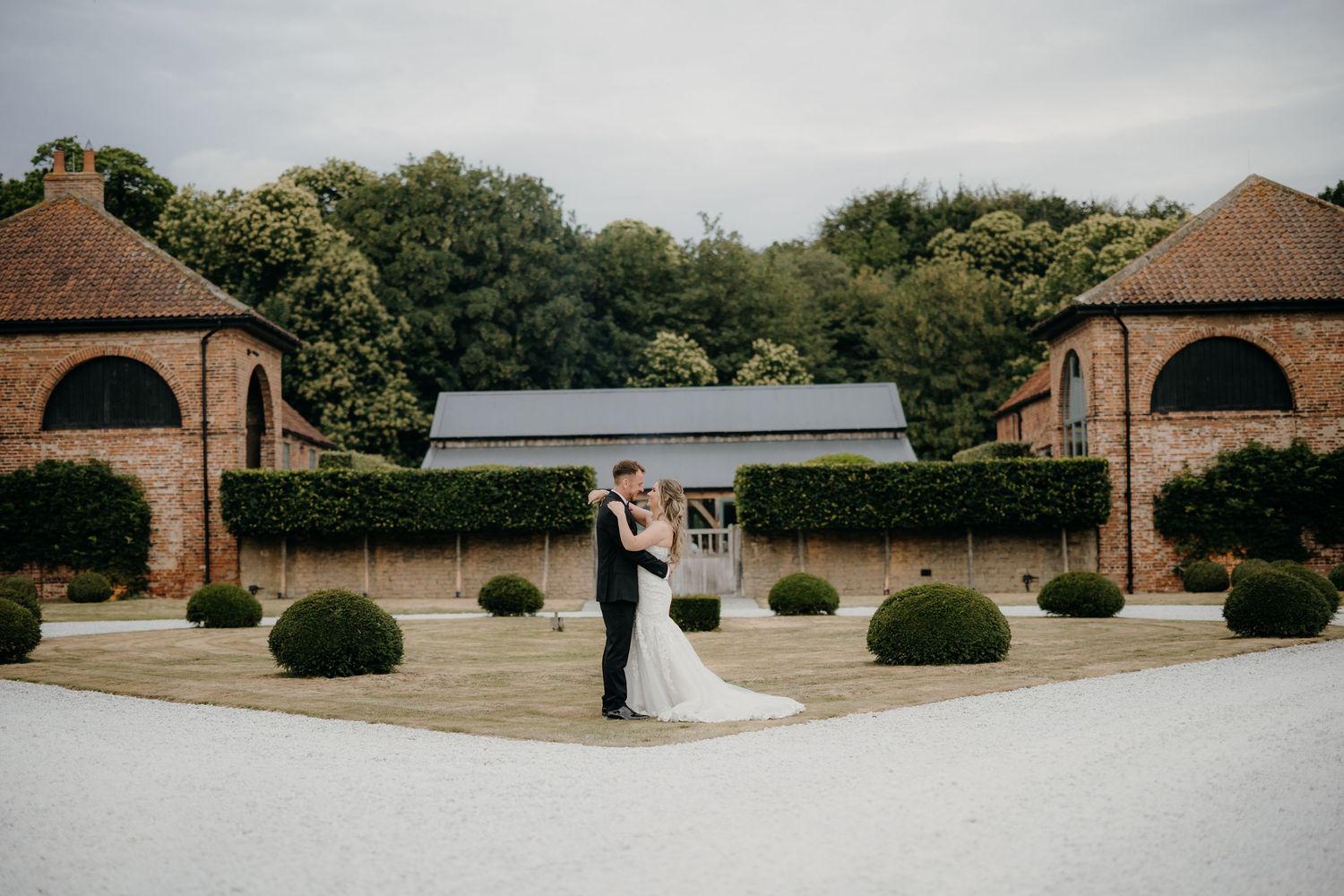 Bride and groom share a romantic embrace in a stunning courtyard venue with manicured hedges and brick buildings.