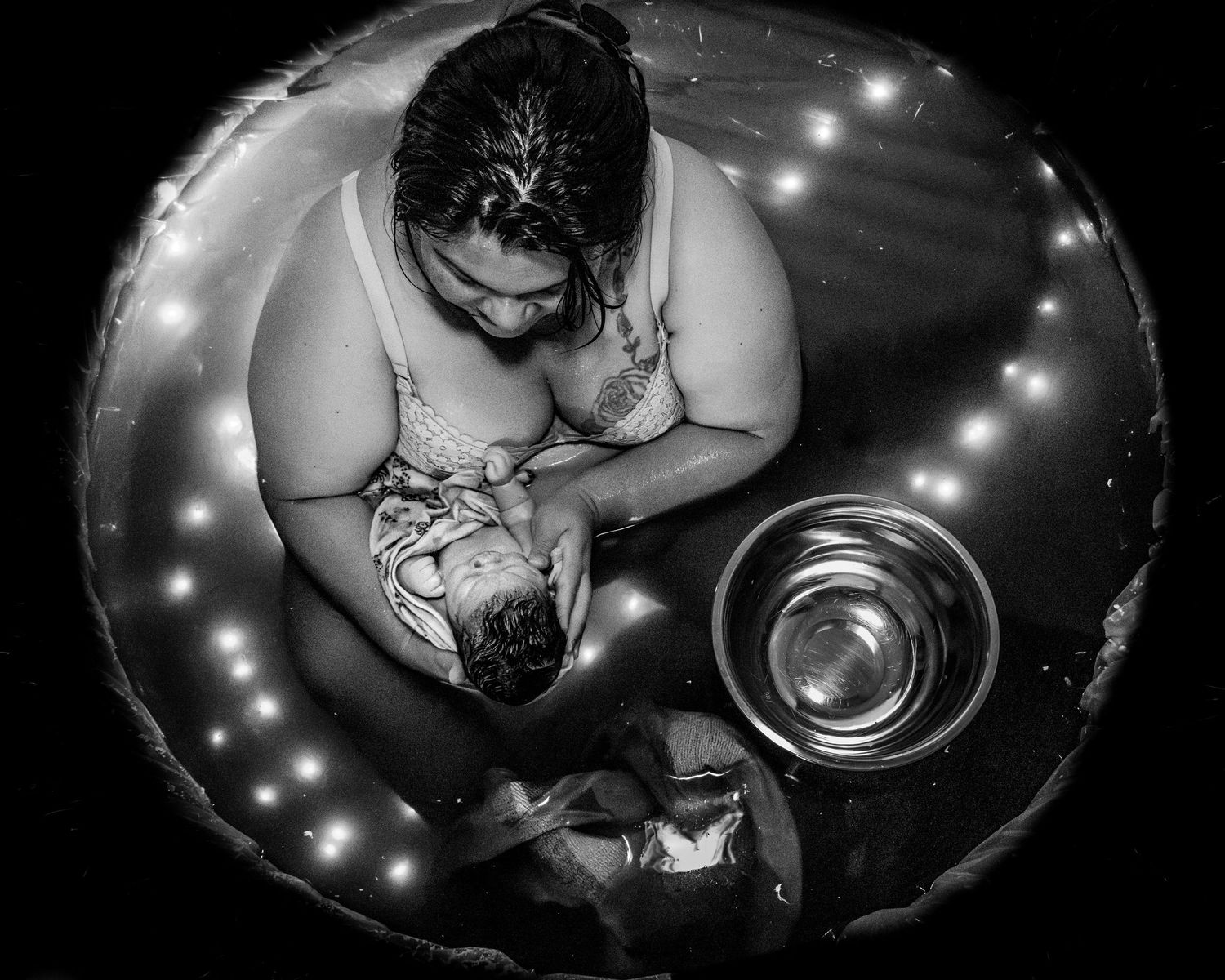 Black and white overhead shot of a mother breastfeeding her newborn in a water birth tub with soft glowing lights.