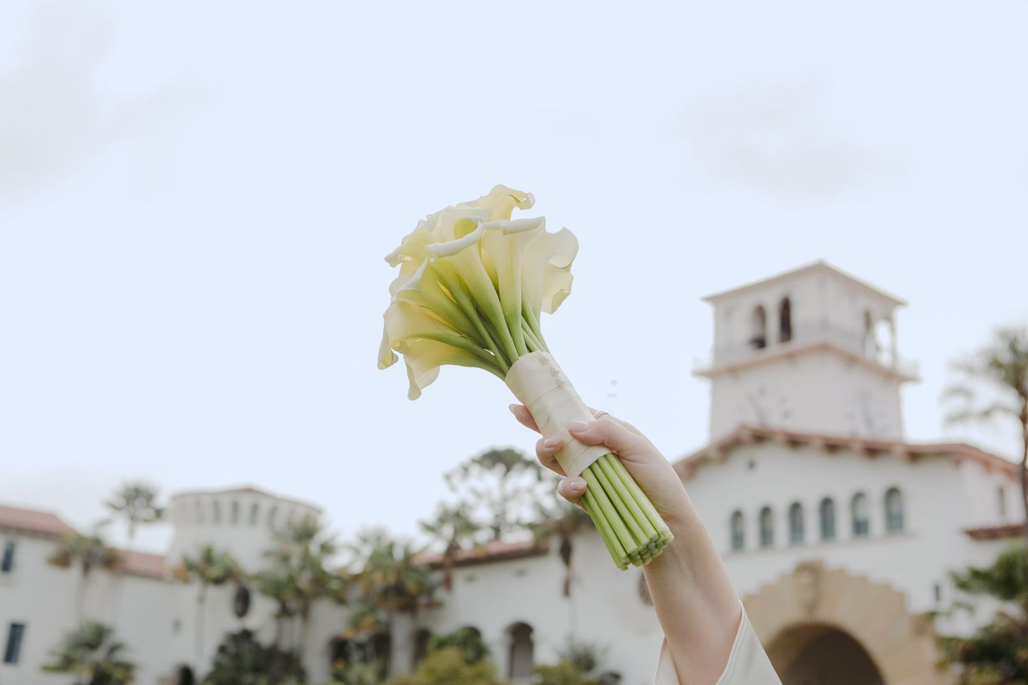 Hands holding a white calla lily bouquet aloft in front of a Spanish-style building with palm trees.