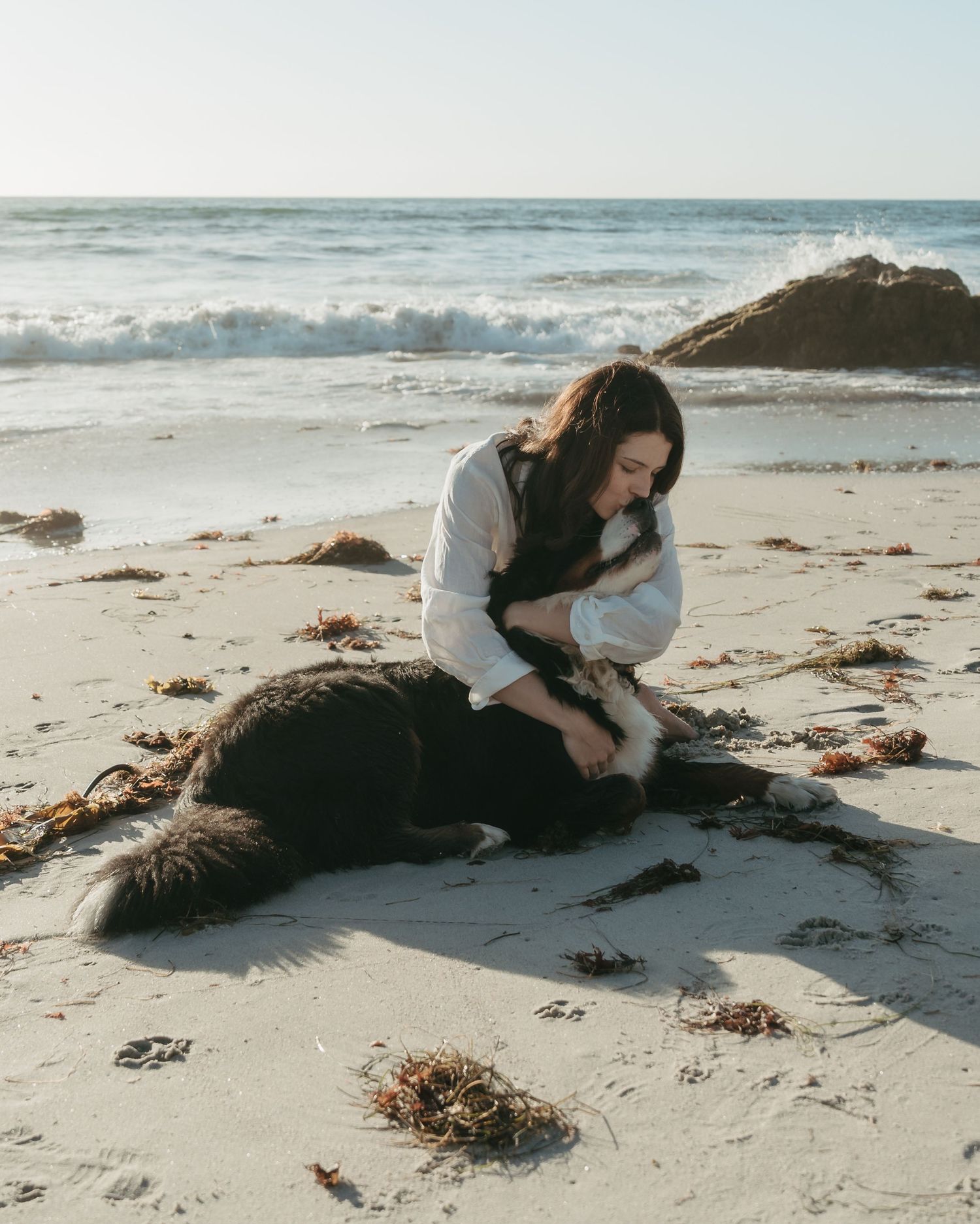 Woman in white shirt cuddling a black and white dog on a sandy beach with ocean waves in the background.