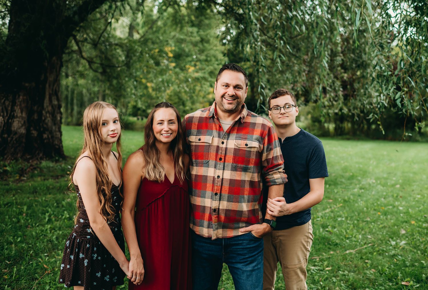 Family of four smiling outdoors in a lush green park, posing together for a portrait photo.