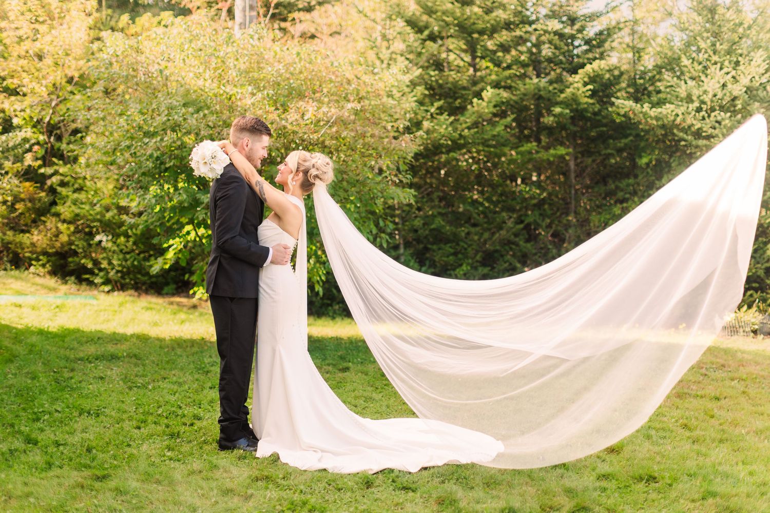 Bride and groom share a romantic kiss outdoors with elegant veil flowing in the wind against lush green trees.