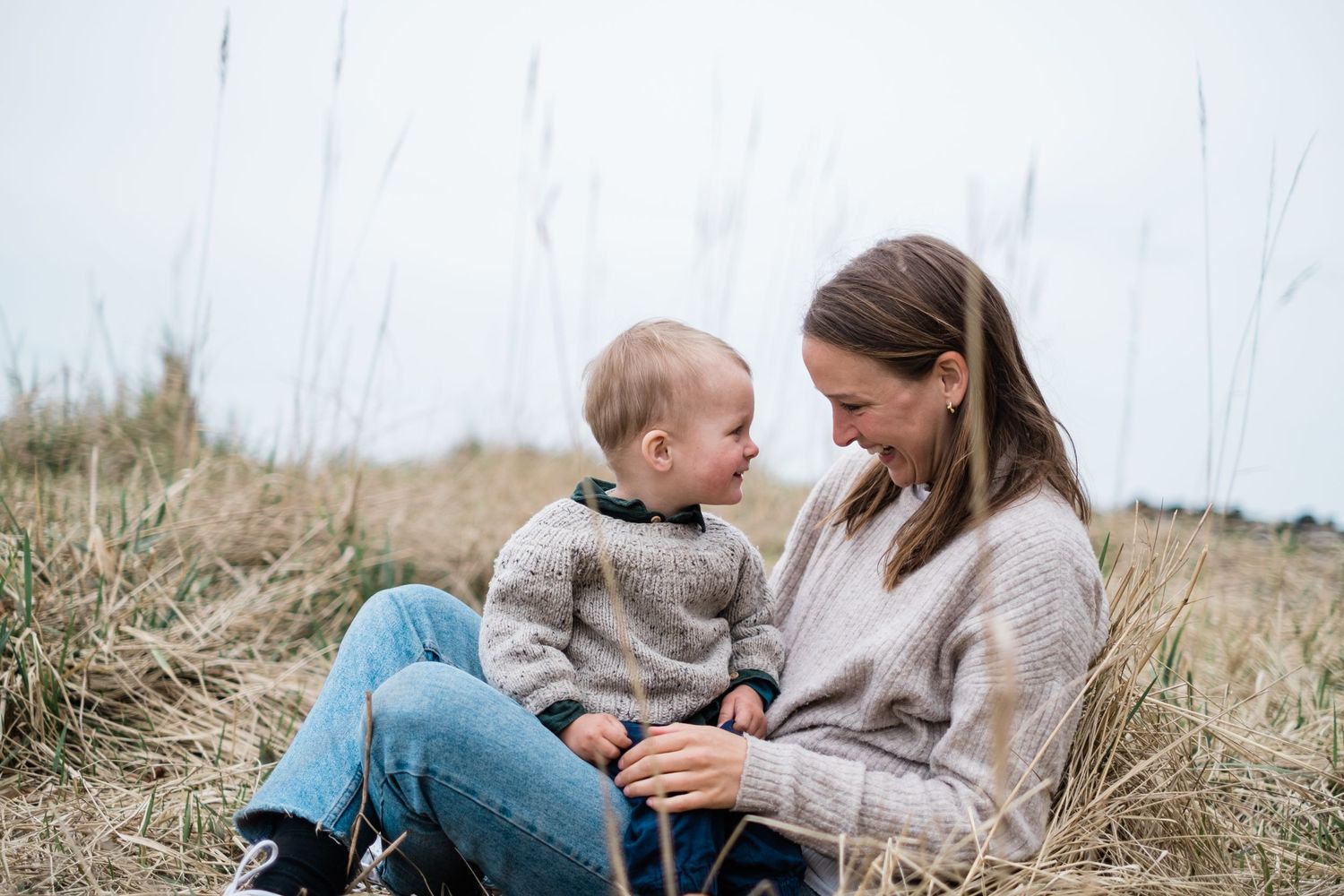 Parent and child share tender moment sitting in tall grass by the coast.