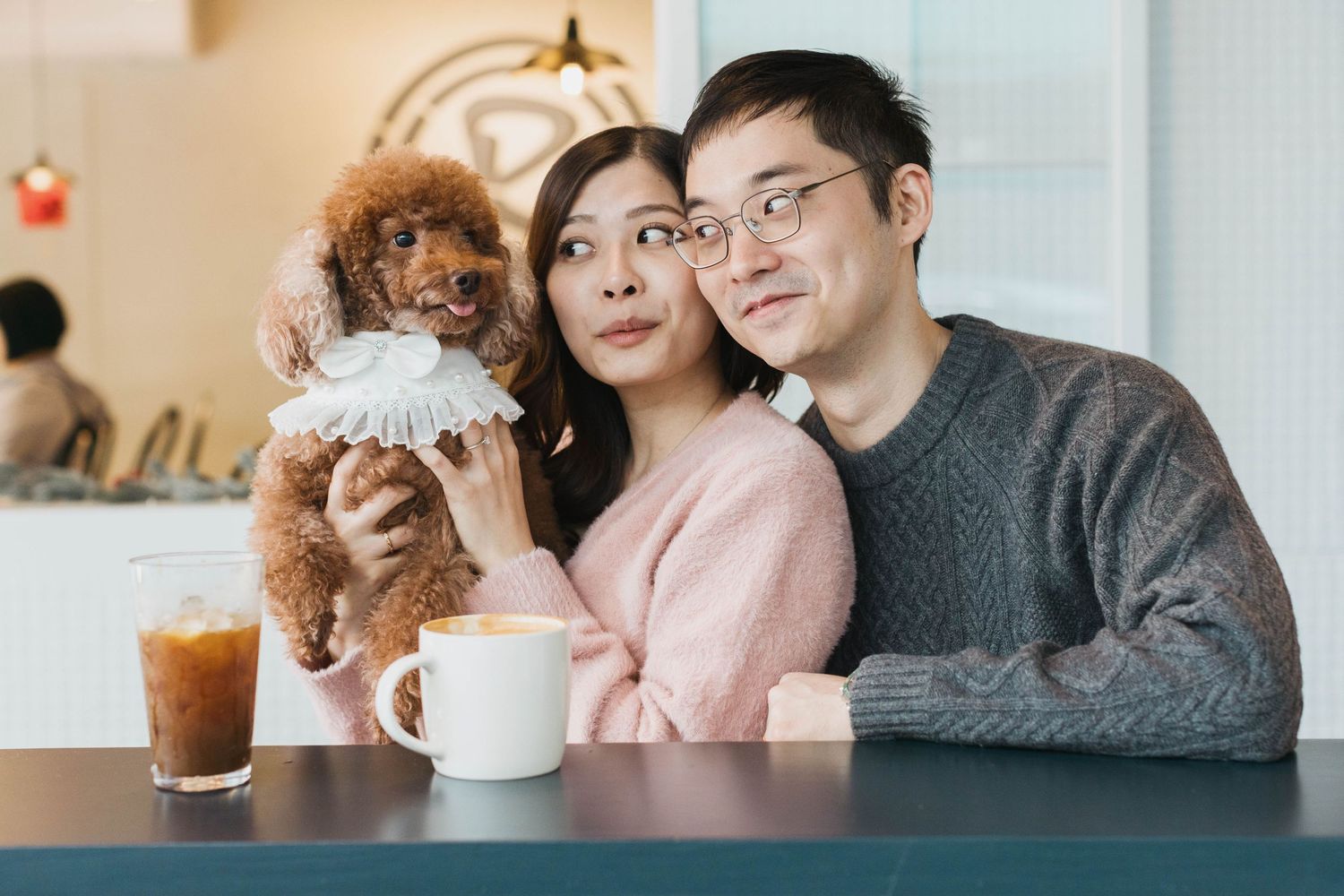 A smiling couple poses with their brown toy poodle at a cozy cafe.