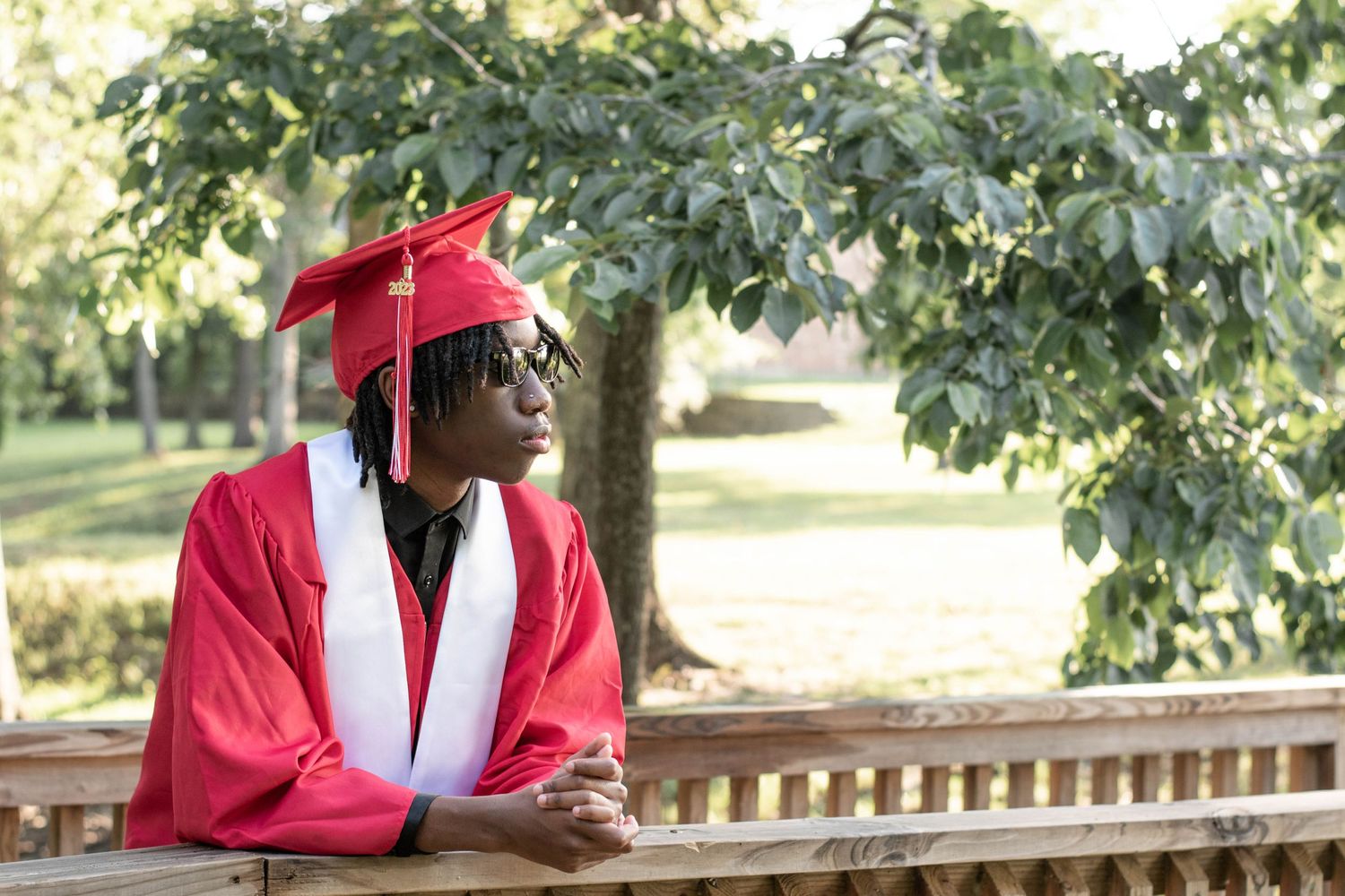 Graduate in bright red cap and gown sitting thoughtfully on a bench in a sunny outdoor setting.
