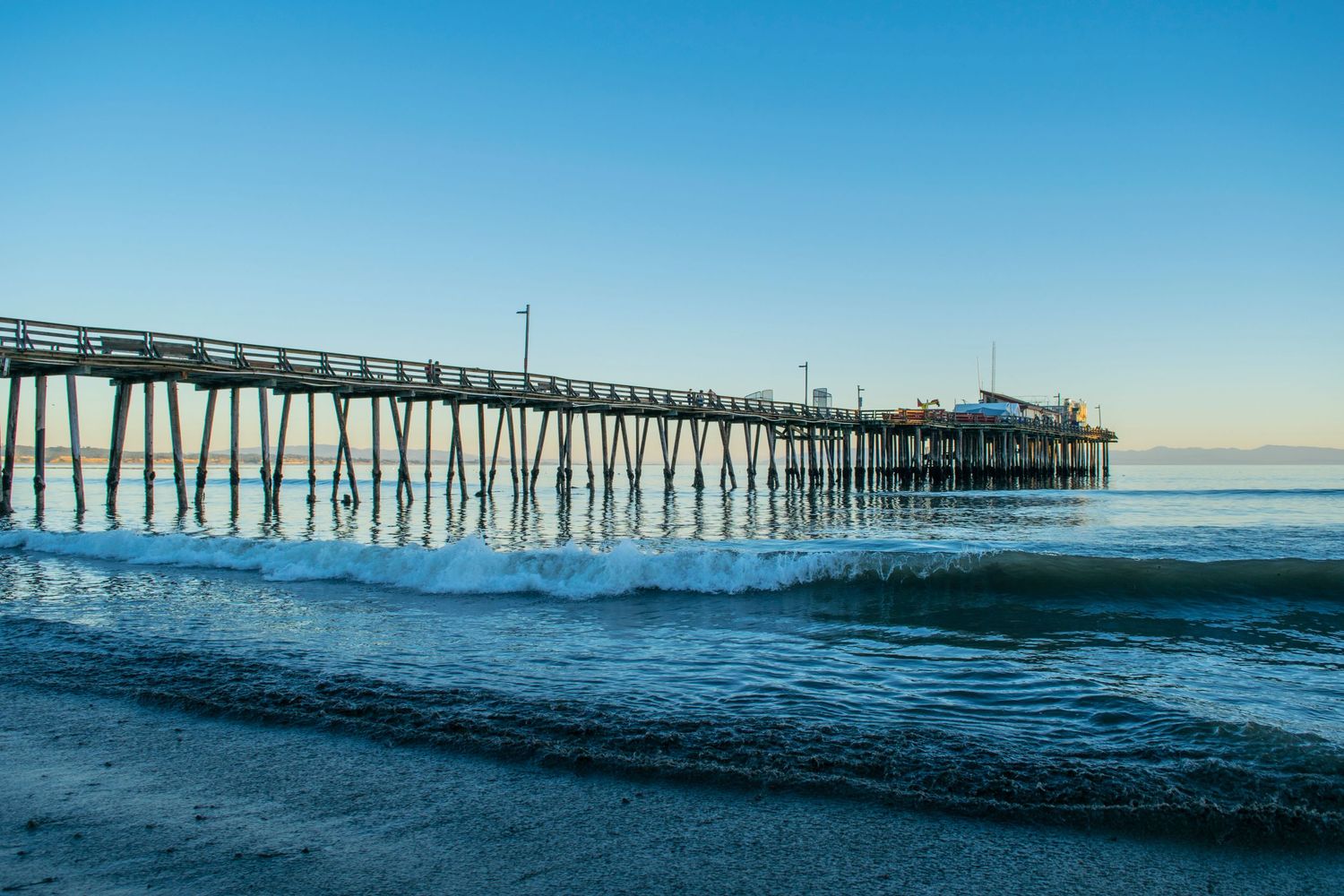 Wooden fishing pier stretches into calm ocean waters under clear blue sky.
