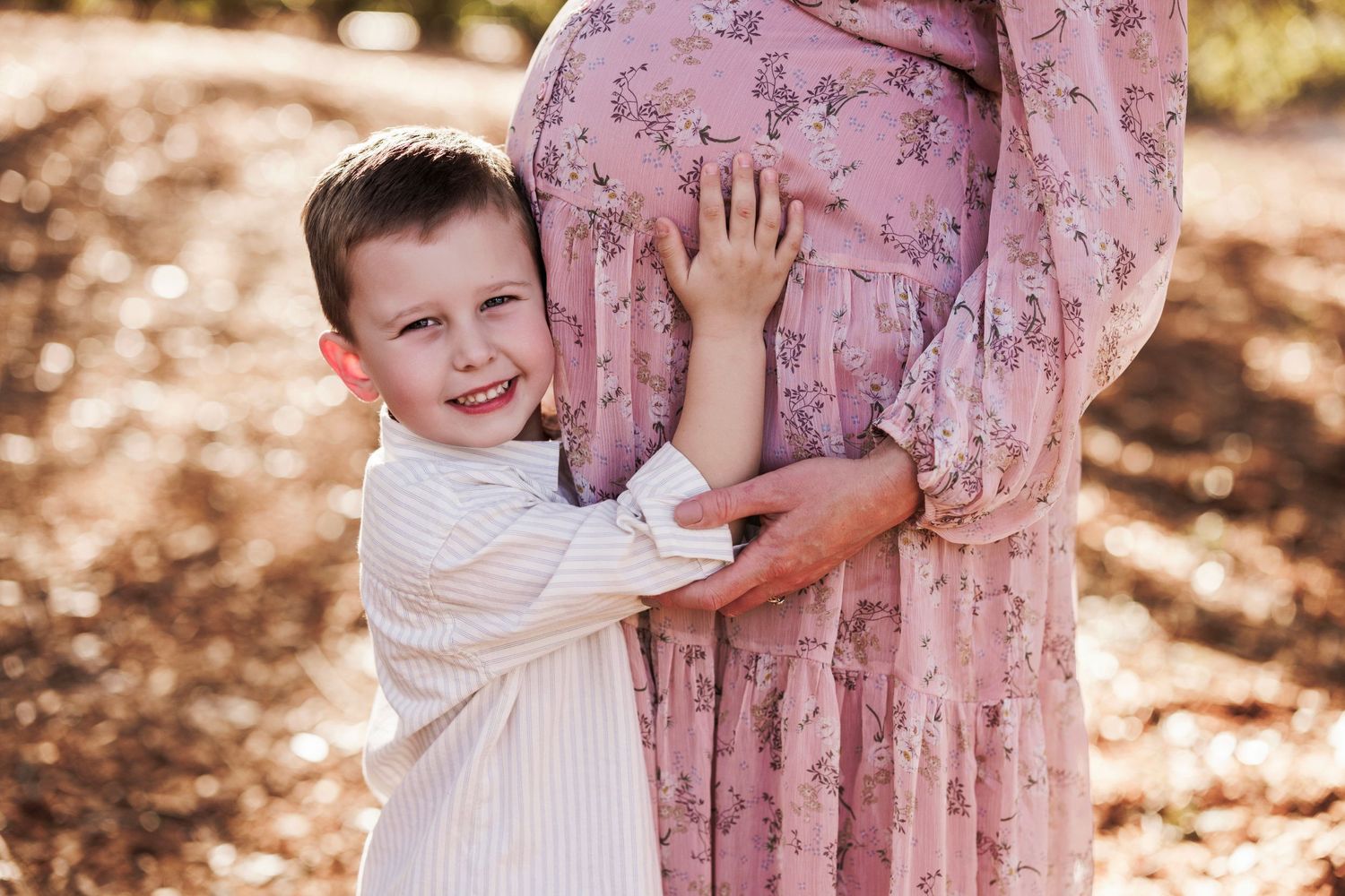A child wearing a white dress embraces a person in a floral pink dress outdoors in dappled sunlight.