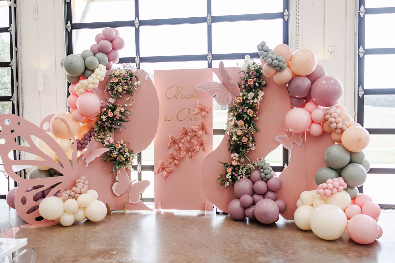 Elegant balloon arch installation in pink and grey tones with floral accents and decorative lights against an industrial window.