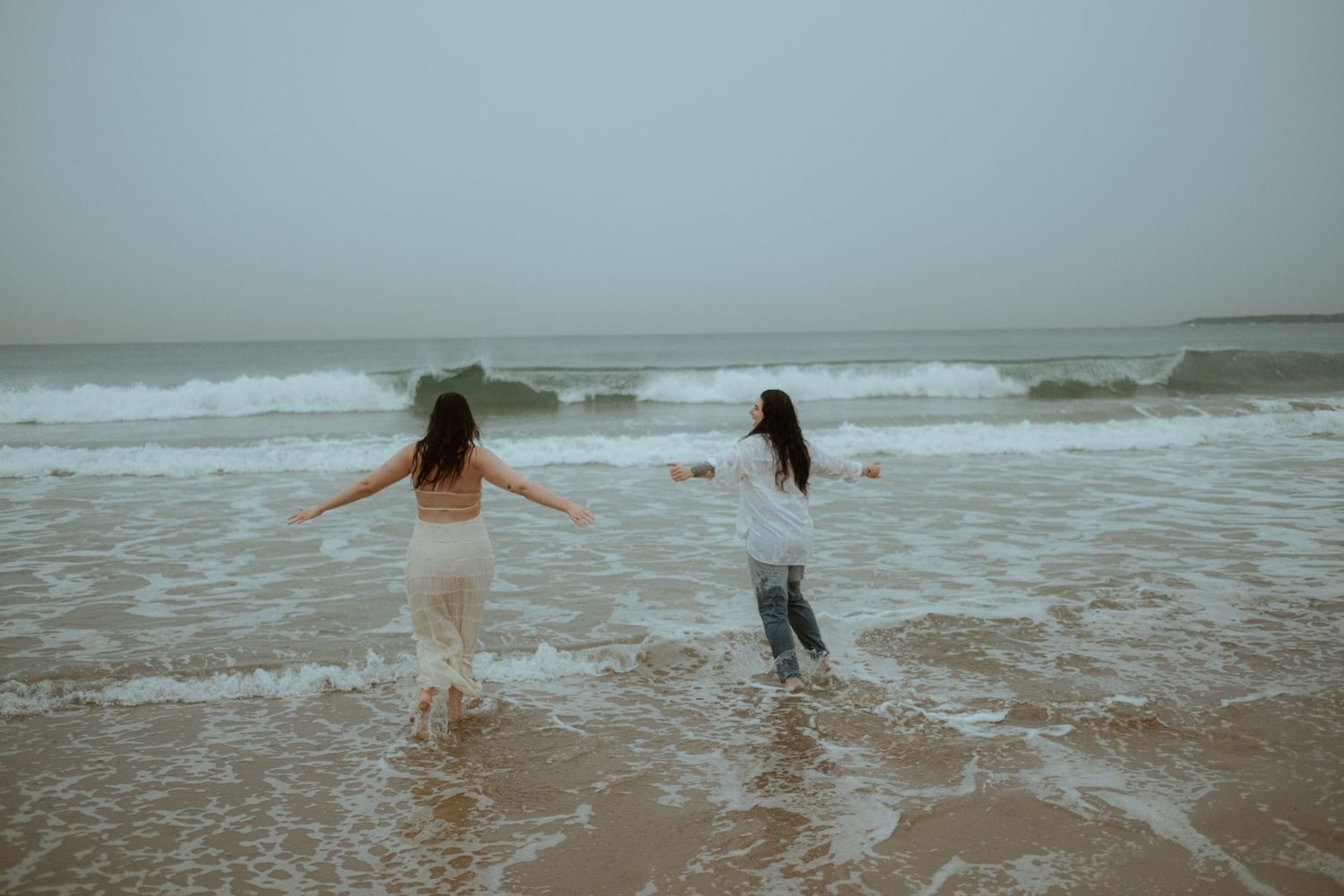 Two people share an intimate moment while standing together on a misty beach as waves roll in along the shoreline.