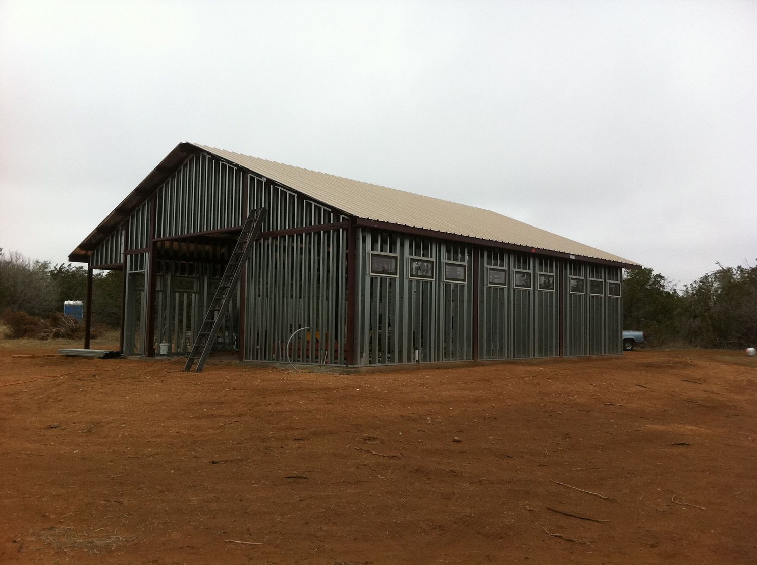 Large green metal barn structure with vertical siding stands in an open dirt field.