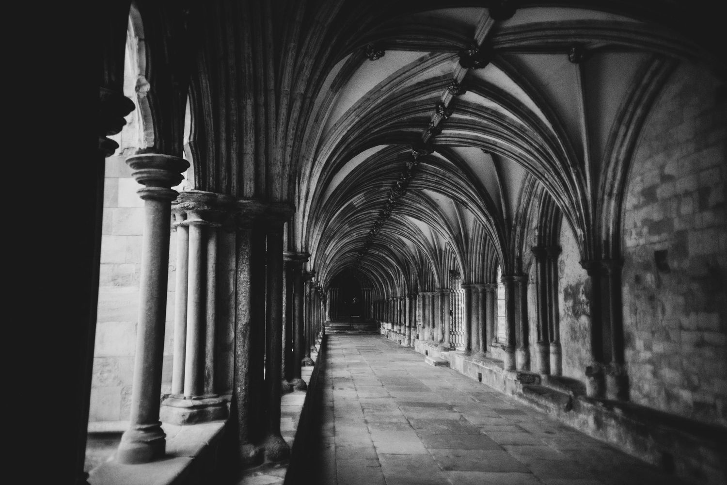 A black and white photograph of a Gothic cathedral cloister with vaulted arches and stone columns creating a long corridor.