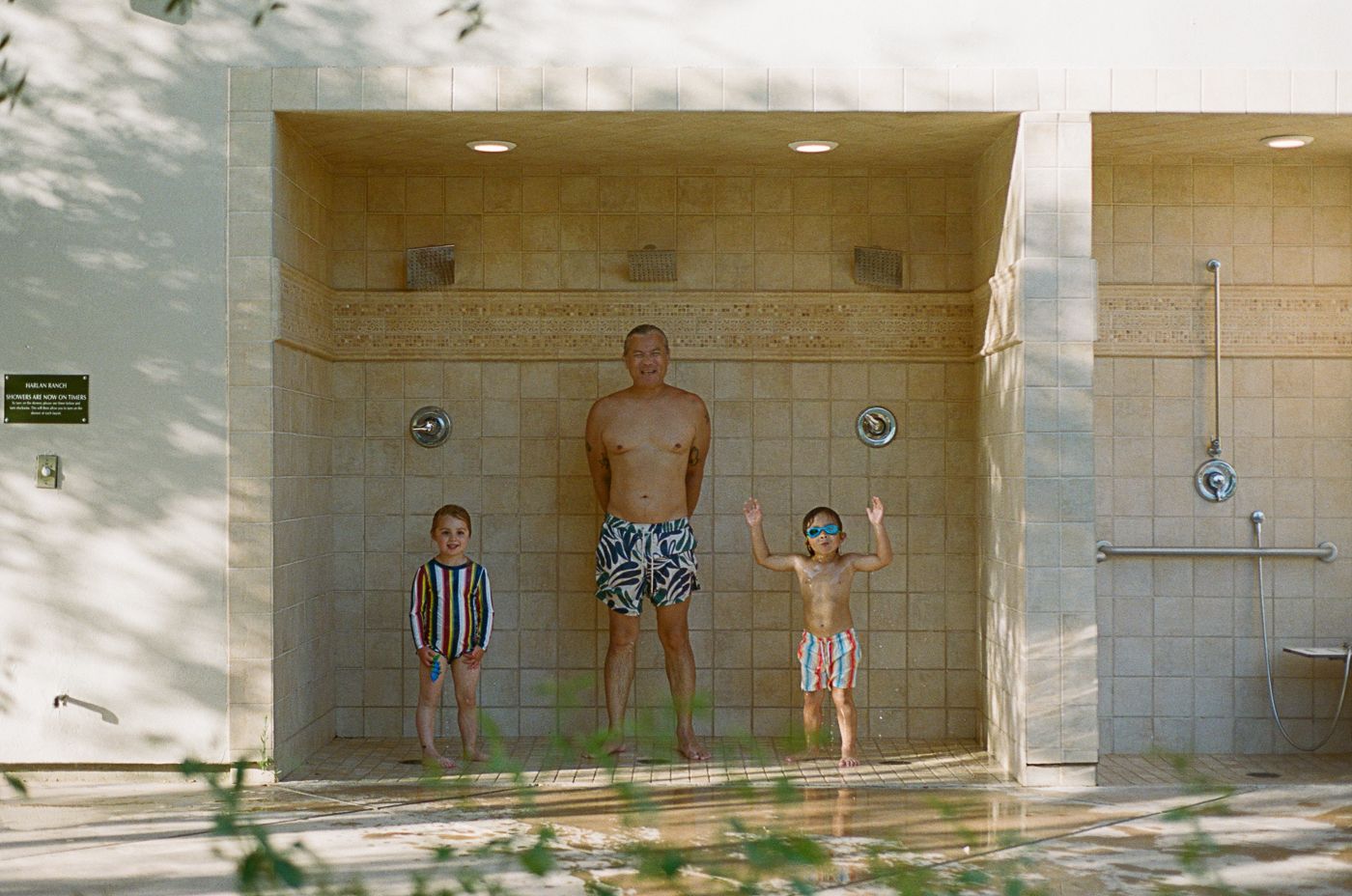 Three figures standing against a tiled wall at what appears to be a beach shower or pool facility.