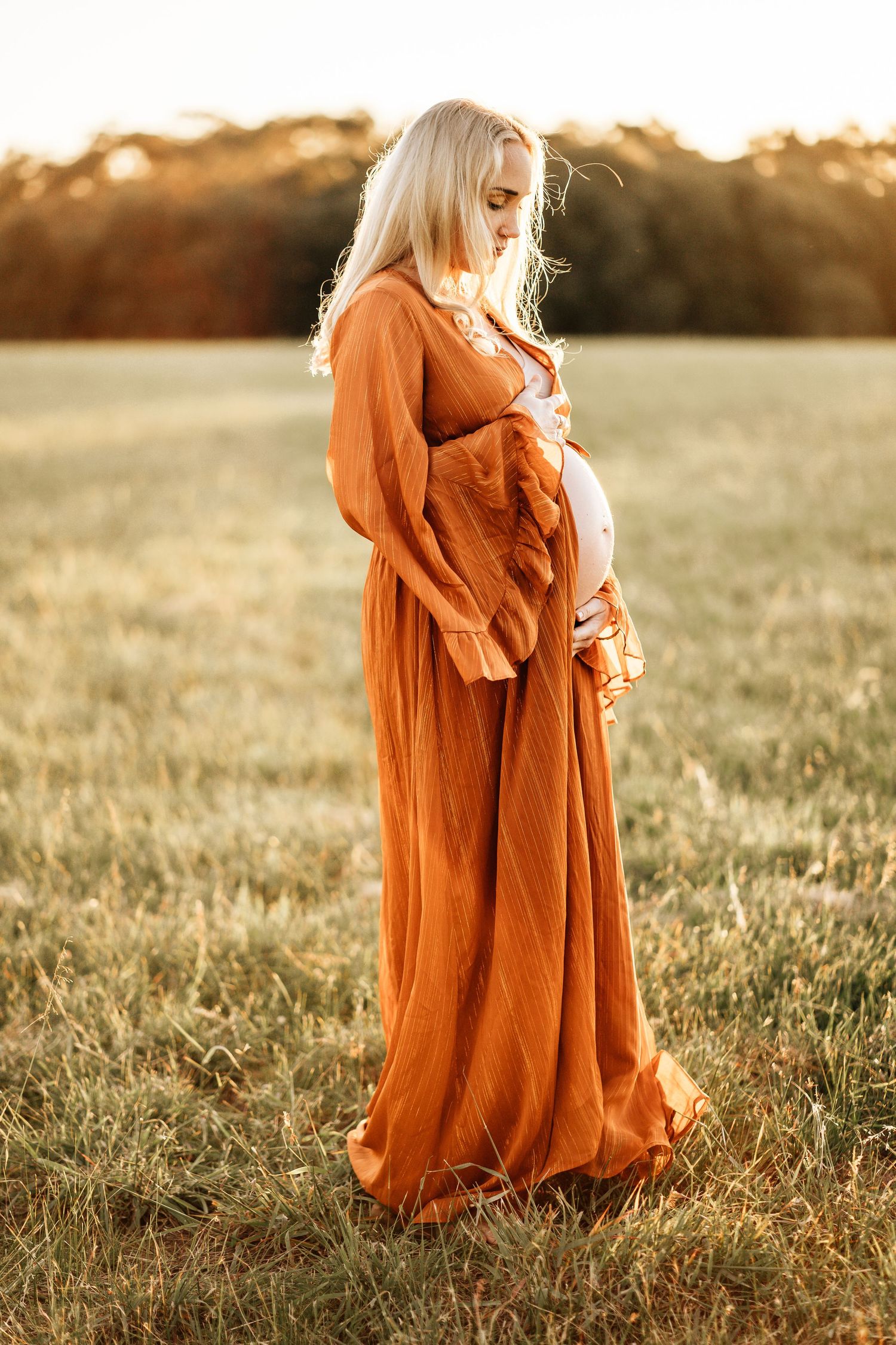 Pregnant woman in flowing rust-colored dress stands in golden field during sunset.