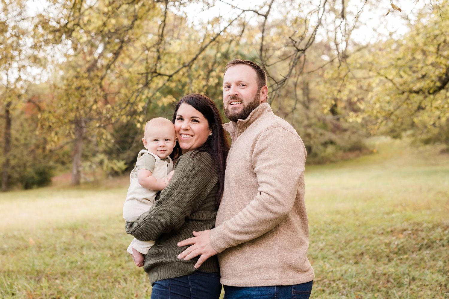 Happy family of three posing outdoors in autumn park with colorful fall foliage.