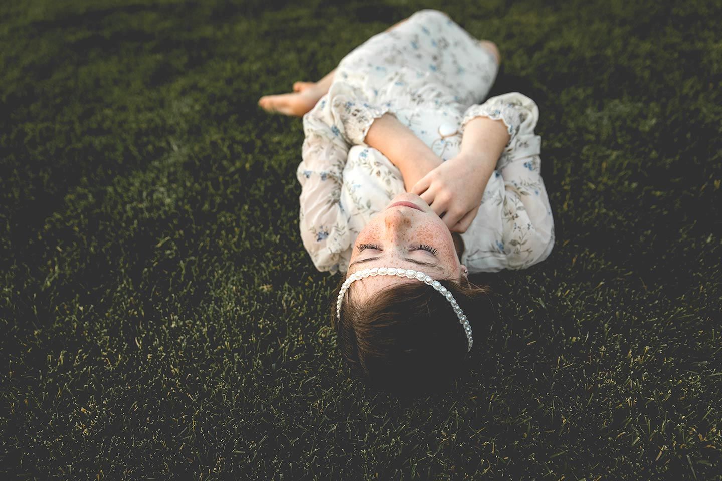 tween girl laying in the grass with a flower dress on and her hand tucked near her chin