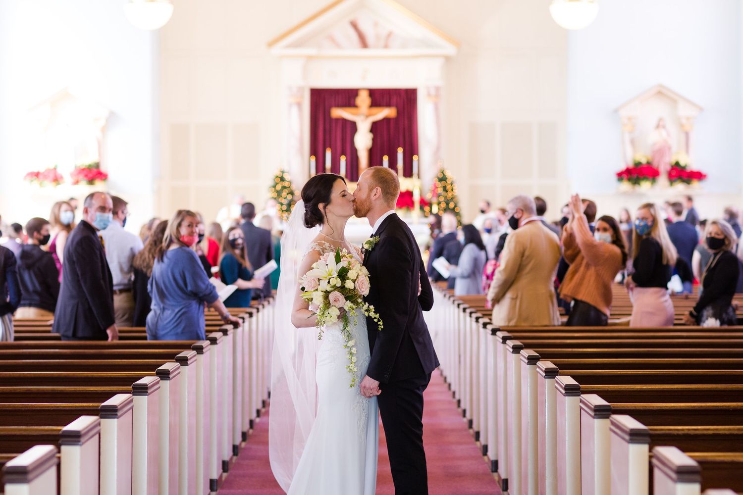 A wedding ceremony in progress inside a traditional church with white pews and red poinsettias.