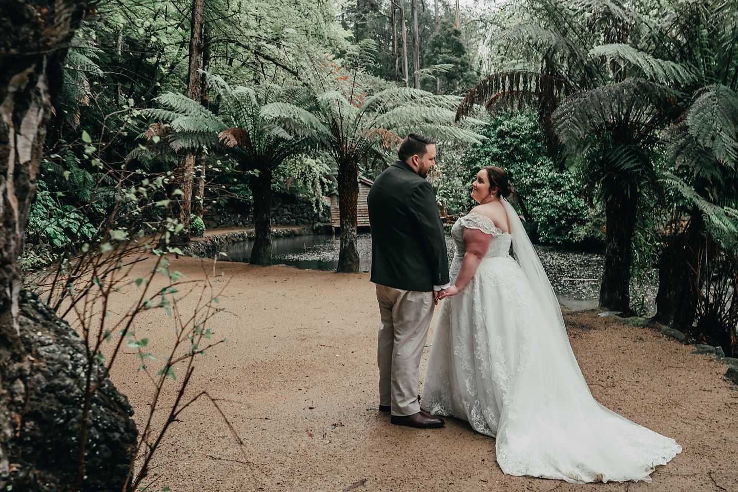 A couple in wedding attire standing on a path surrounded by lush tropical gardens and palm trees.