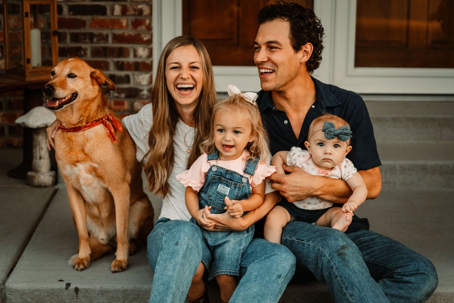 Happy family sitting on front porch steps with their golden retriever dog and two small children.