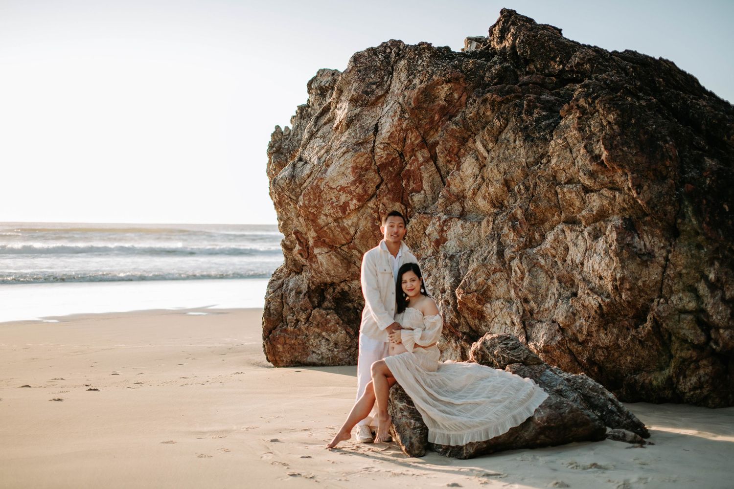 A romantic maternity photoshoot featuring a couple posed against dramatic beach rocks.