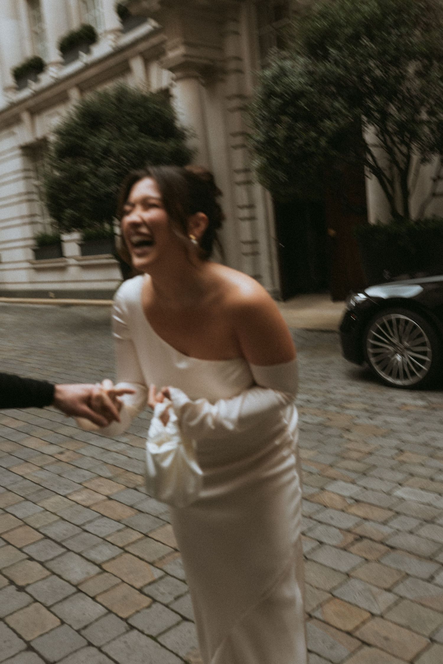 A bride in a fitted white dress laughs joyfully on a cobblestone street.
