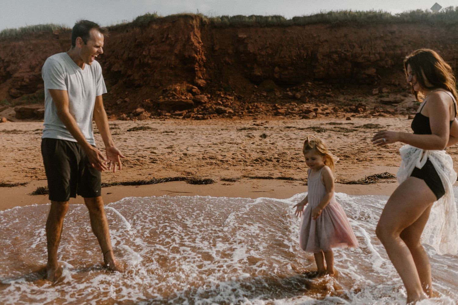 Family plays together at beach with waves splashing around their feet at sunset.