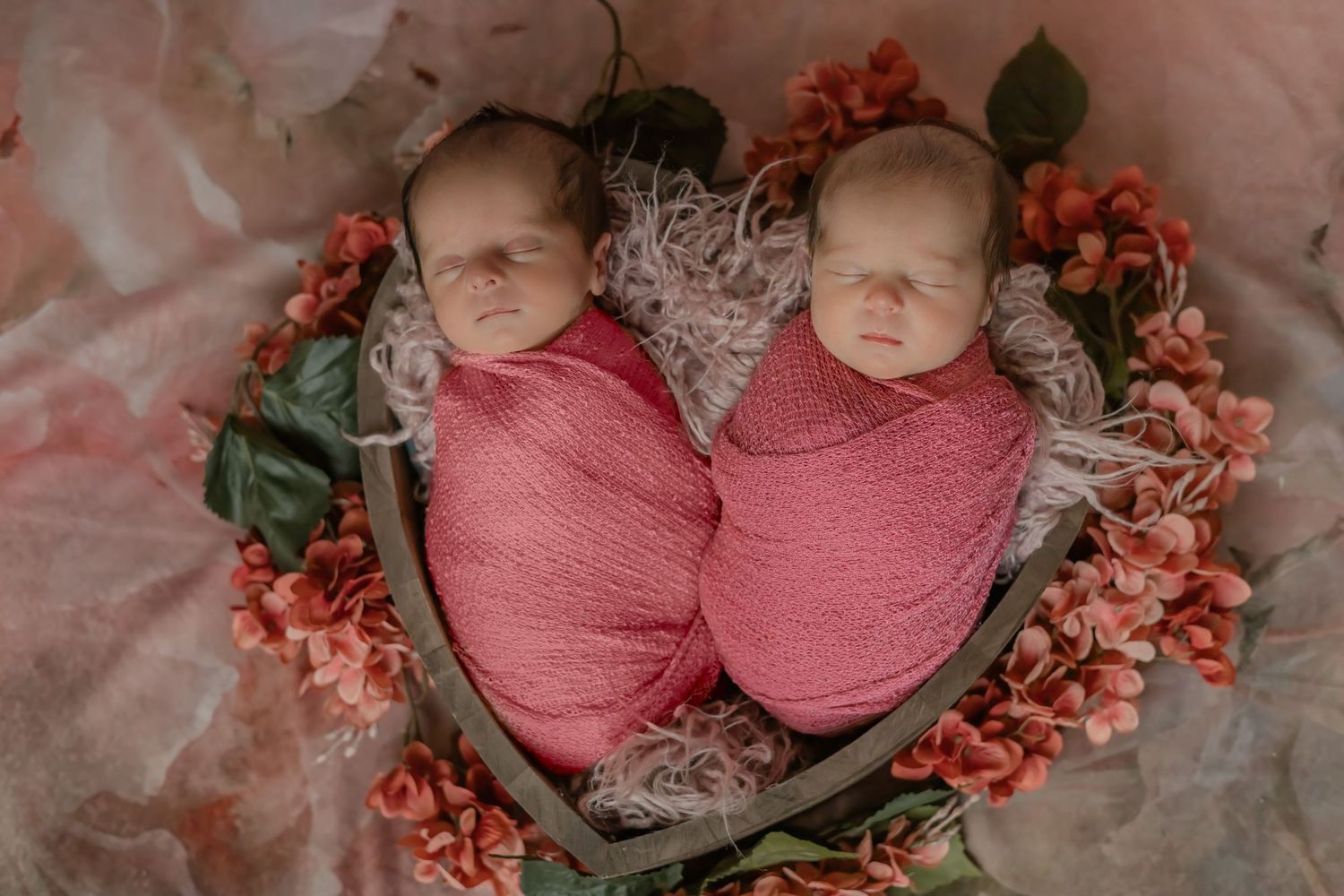 Twin newborns swaddled in pink wraps surrounded by coral flowers.