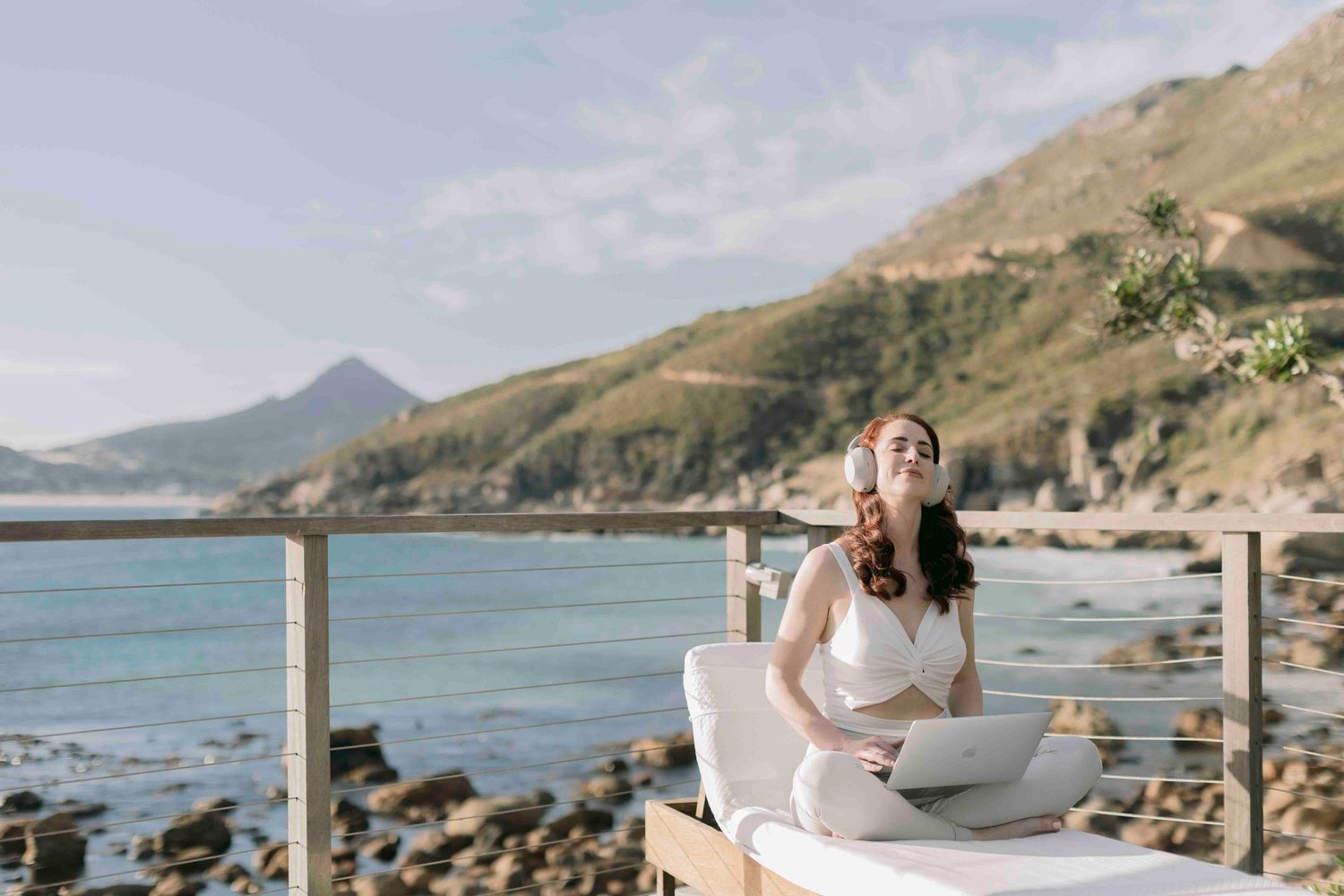 Woman in white outfit sitting on a massage table with a laptop, enjoying coastal mountain views.