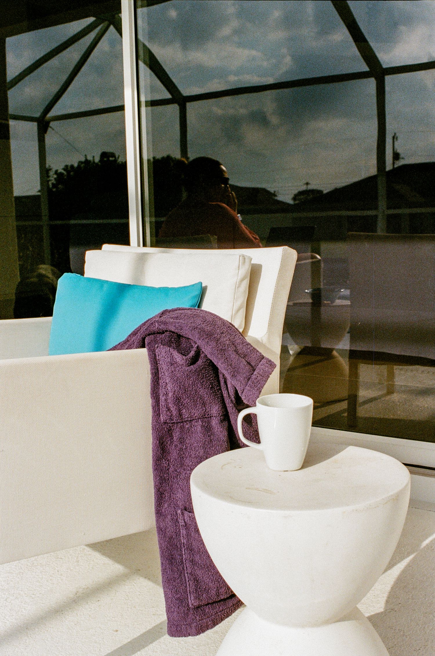 A relaxing sunroom setup with white furniture, turquoise pillow, purple towel and coffee mug on a white side table.