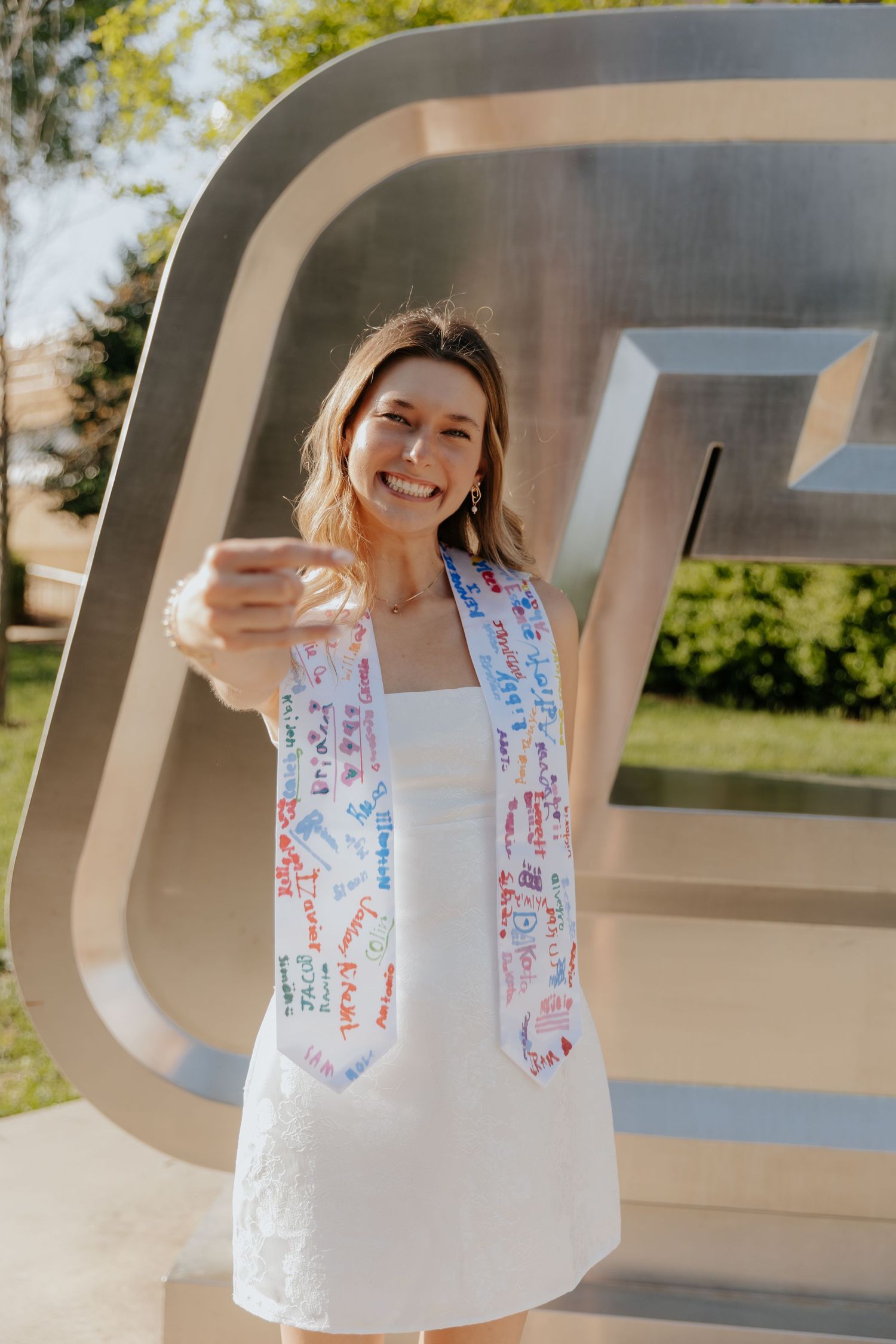 Someone wearing a white dress and colorful scarf poses in front of a large outdoor letter sculpture on a sunny day.
