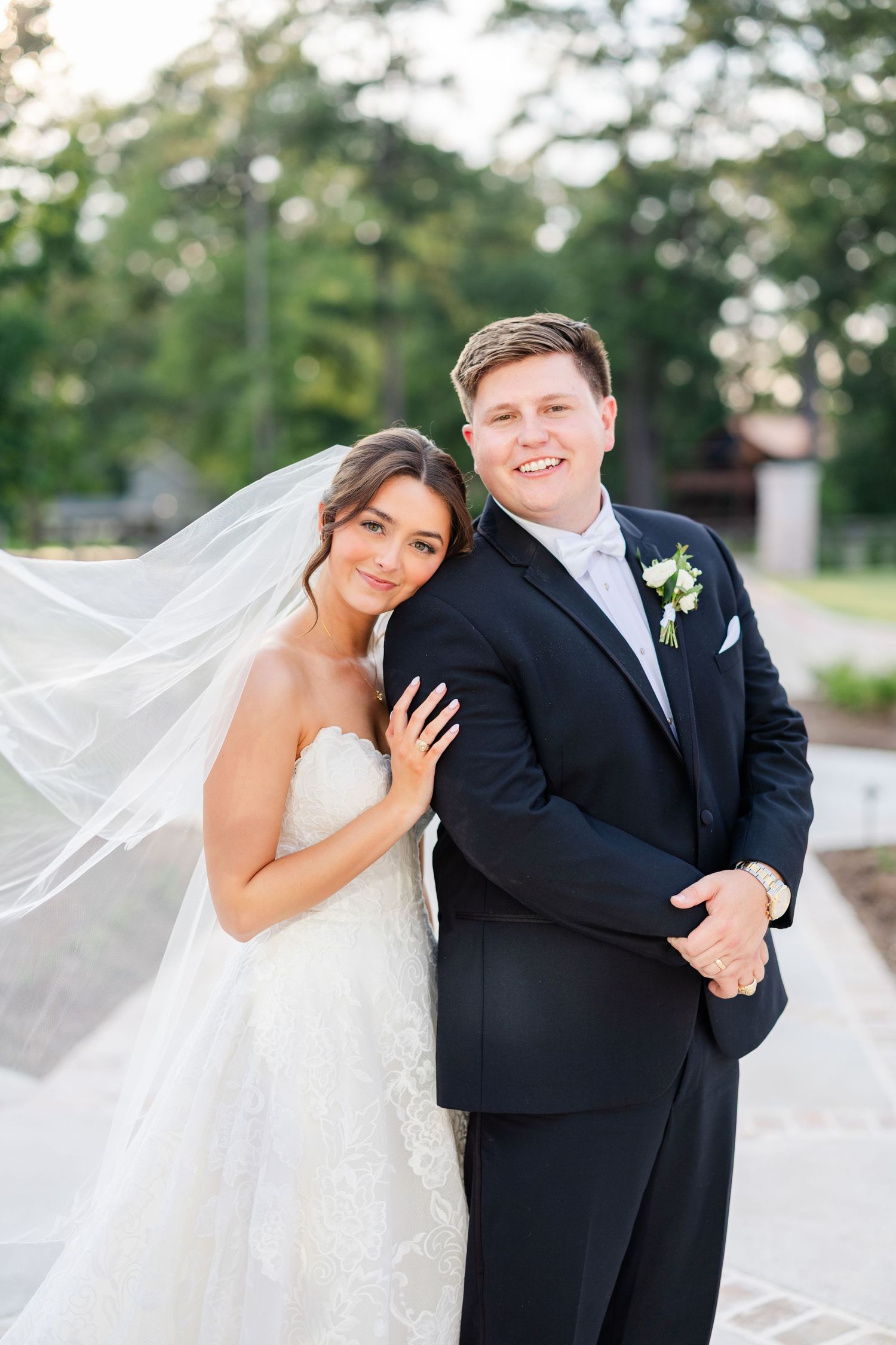 A bride in a strapless white gown and long veil poses with a groom wearing a classic black tuxedo outdoors.