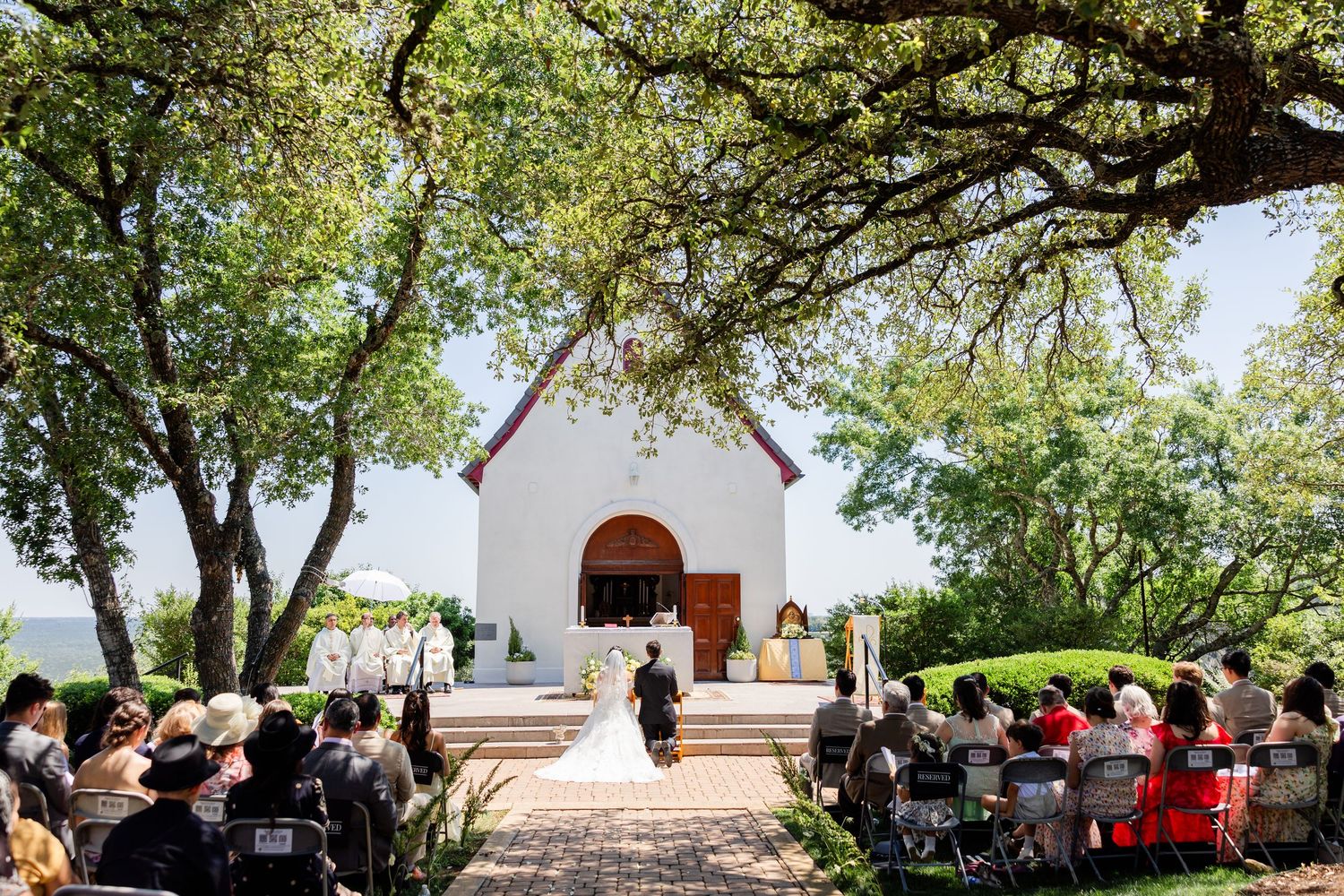 A small white chapel nestled among trees hosts an outdoor wedding ceremony with guests seated on the lawn.