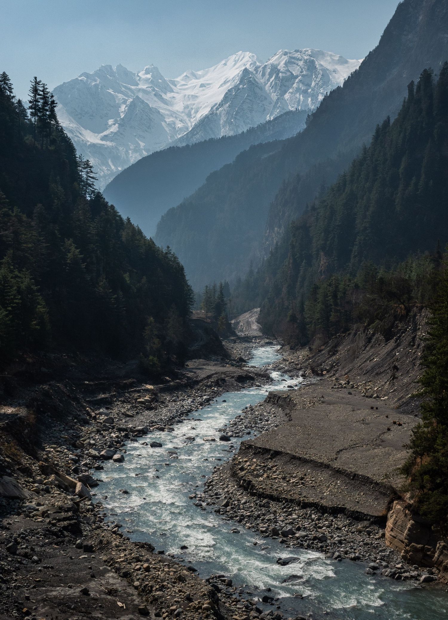 A rushing river winds through a deep mountain valley with snow-capped peaks and evergreen forests in the background.