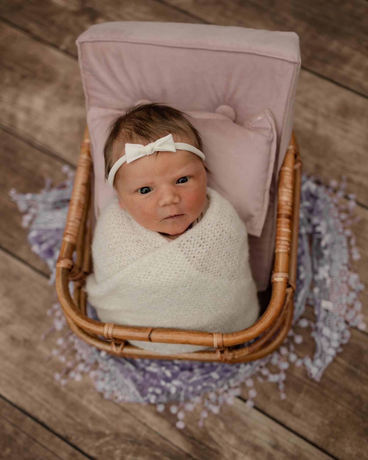 Newborn baby swaddled in white blanket resting in wicker basket on wooden floor.
