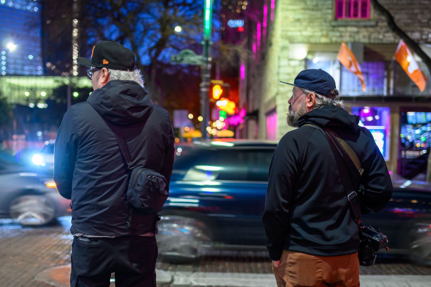 Two men in black jackets standing on city street at night with colorful neon lights in background.