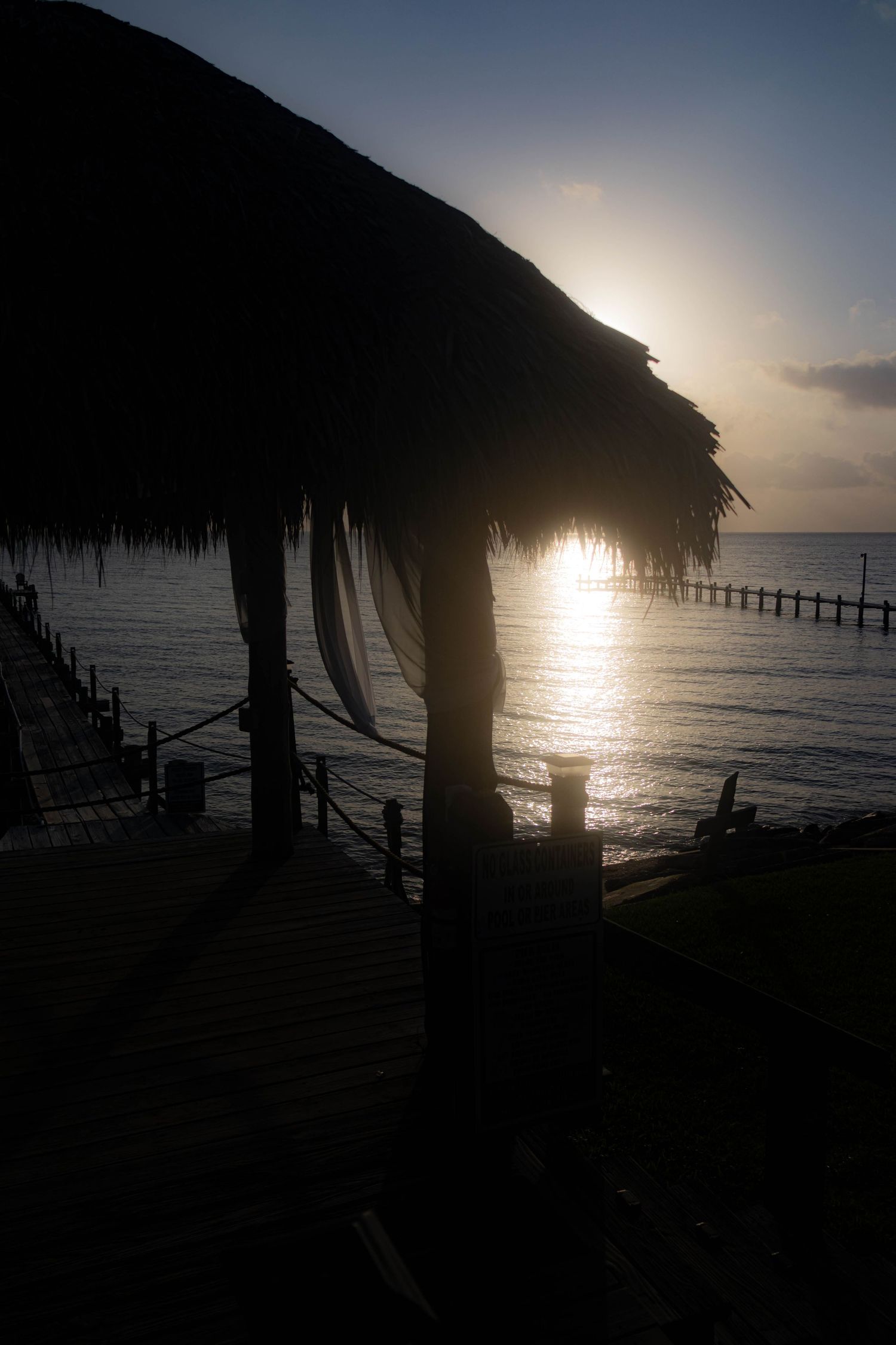 Silhouette of a palm-thatched palapa against a sunset over the ocean with a wooden pier in the distance.