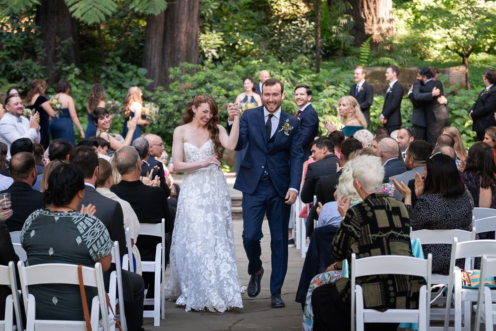 A wedding ceremony takes place in a lush outdoor garden setting with white chairs lined up for guests.