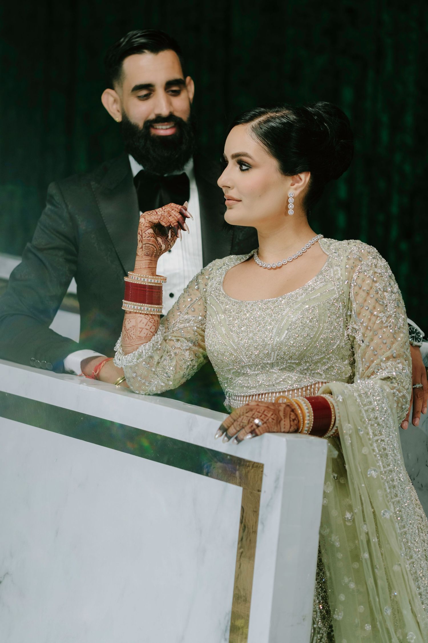 A couple in formal South Asian wedding attire smiles together against a dark green backdrop.