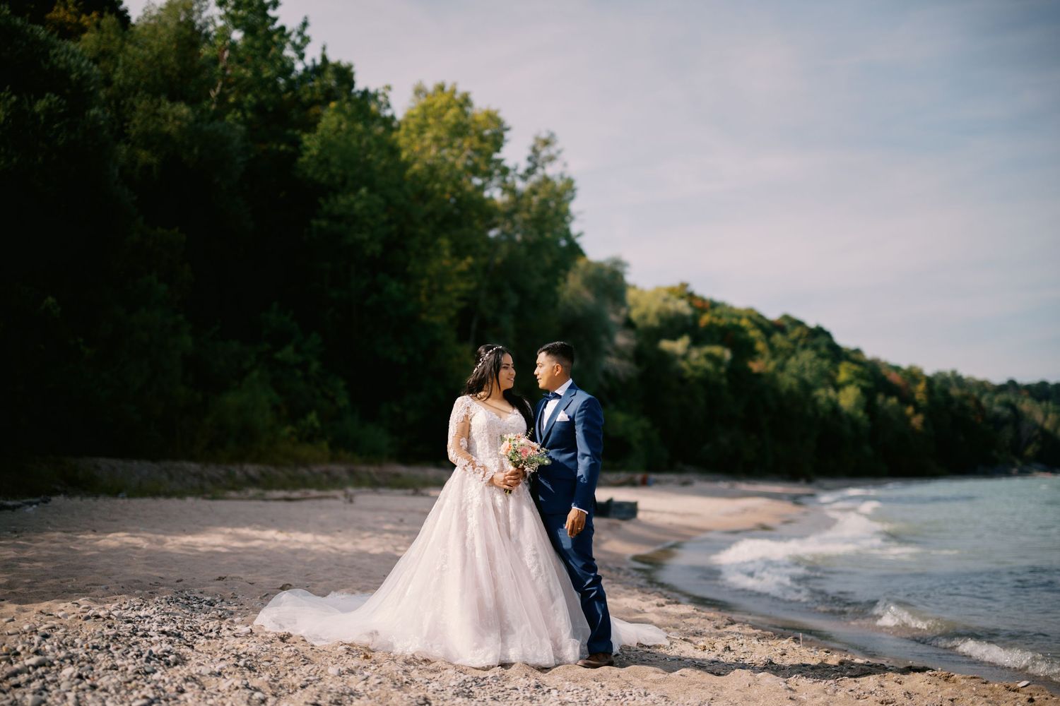 A couple in wedding attire stands lovingly together on a pebble beach shoreline with forested cliffs in the background.